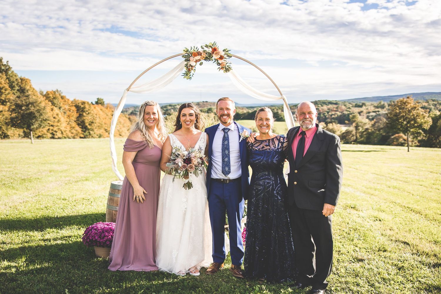 Wedding party poses for group photos under circular arch with mountain landscape backdrop.