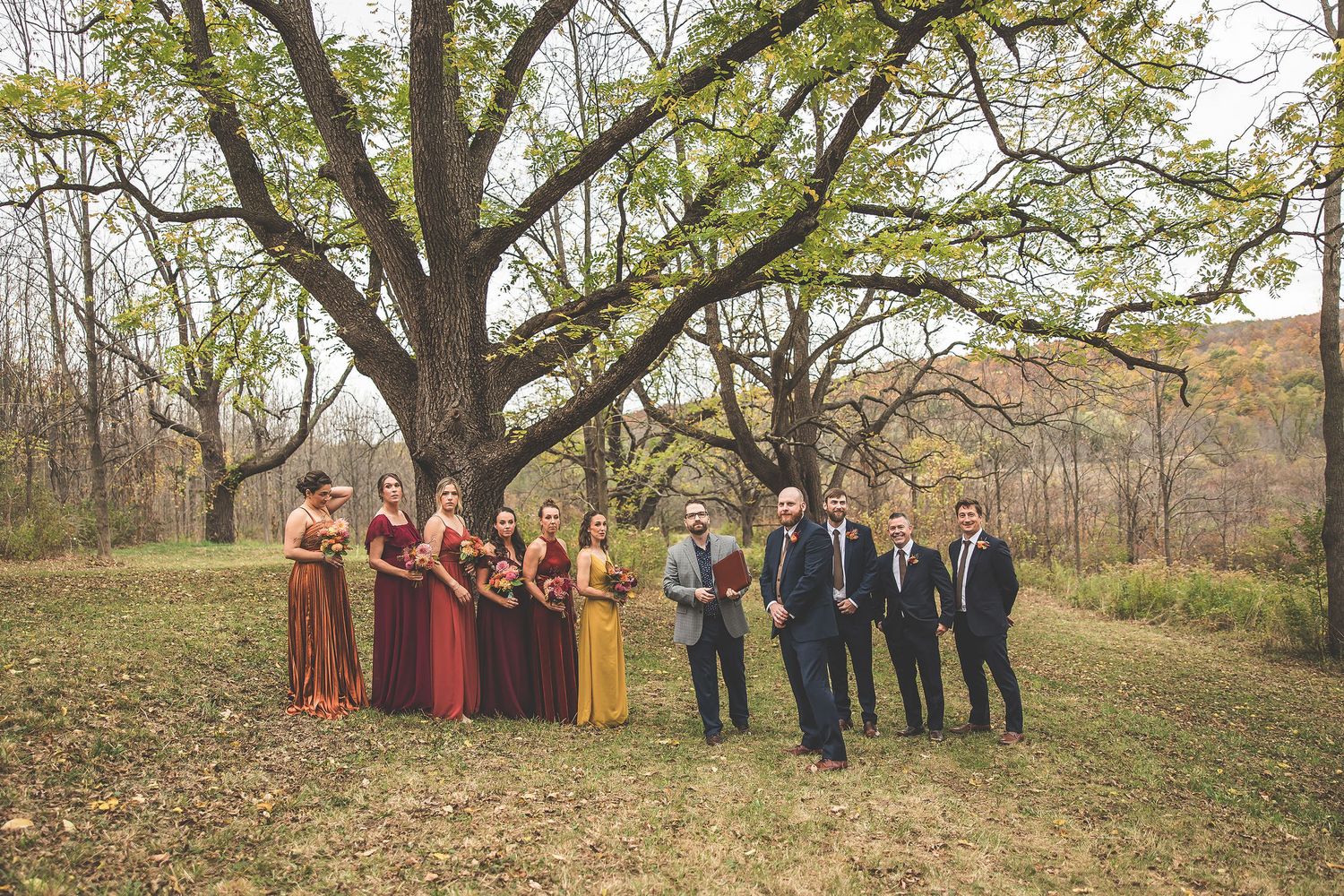 Wedding party arranged in scenic fall landscape with large trees and golden leaves.