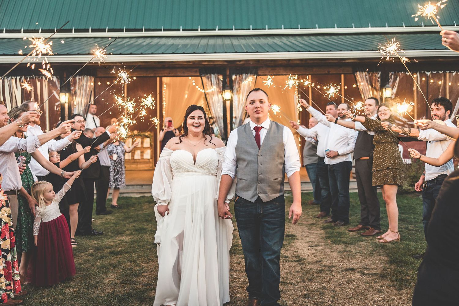 Wedding couple exits through sparkler sendoff line outside barn venue at dusk.