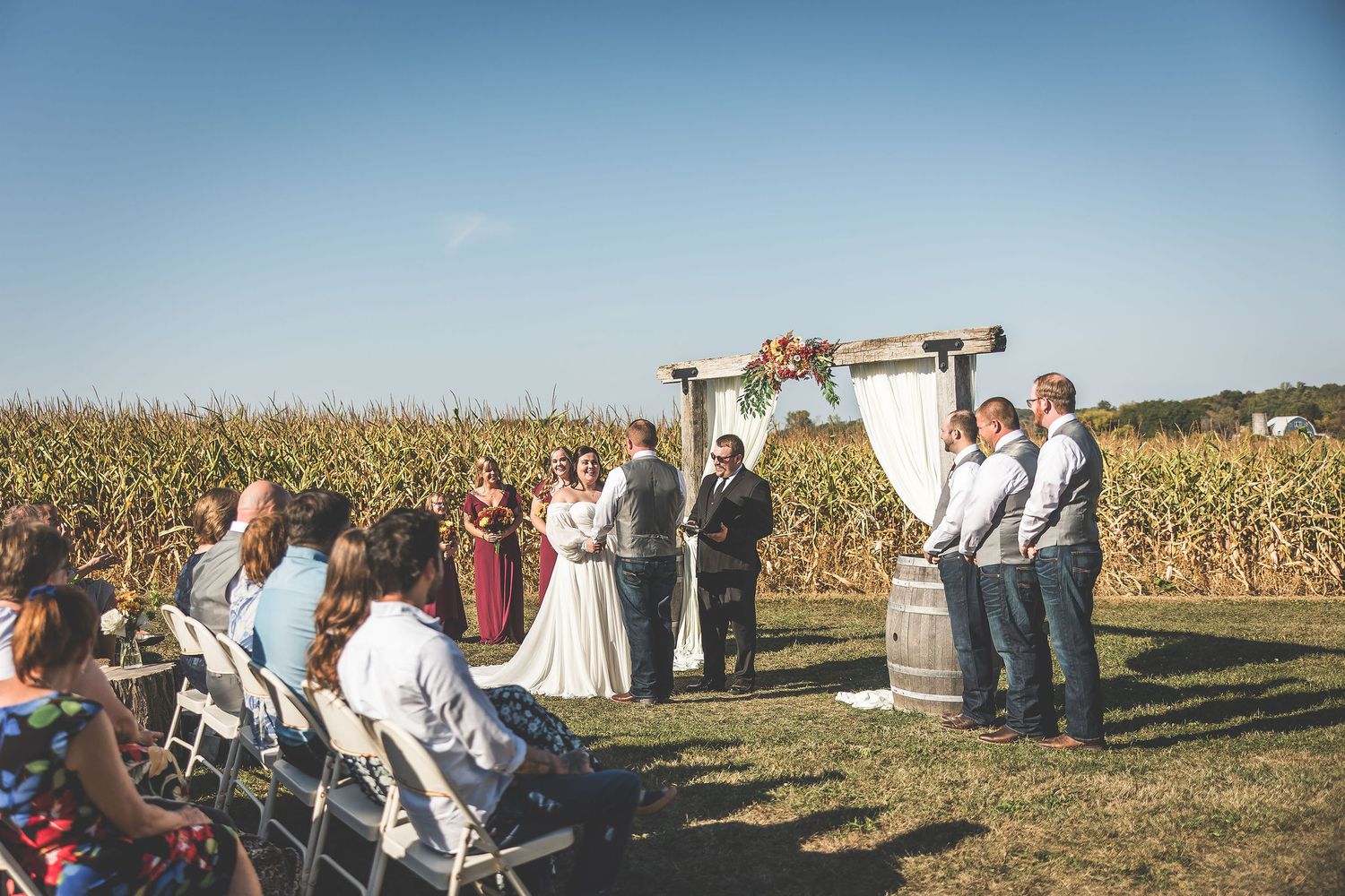 Panoramic view of wedding guests seated outdoors with cornfield and blue sky in the background.