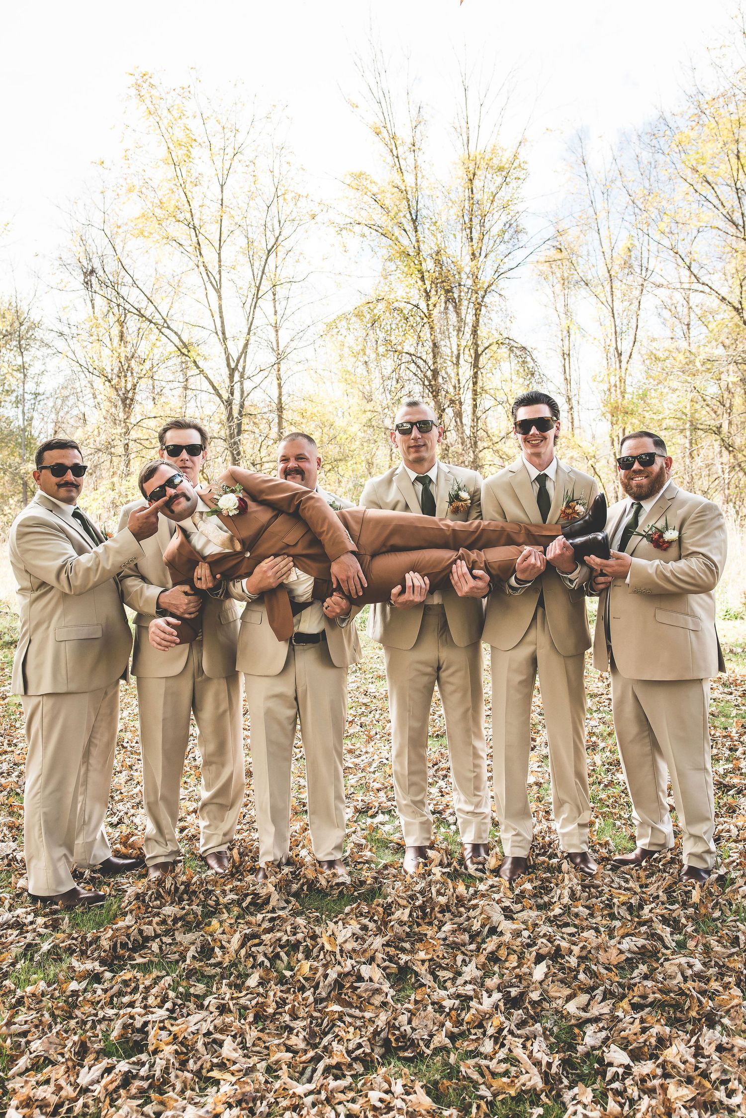 Groomsmen in beige suits pose playfully with sunglasses in autumn leaves during outdoor wedding photoshoot.