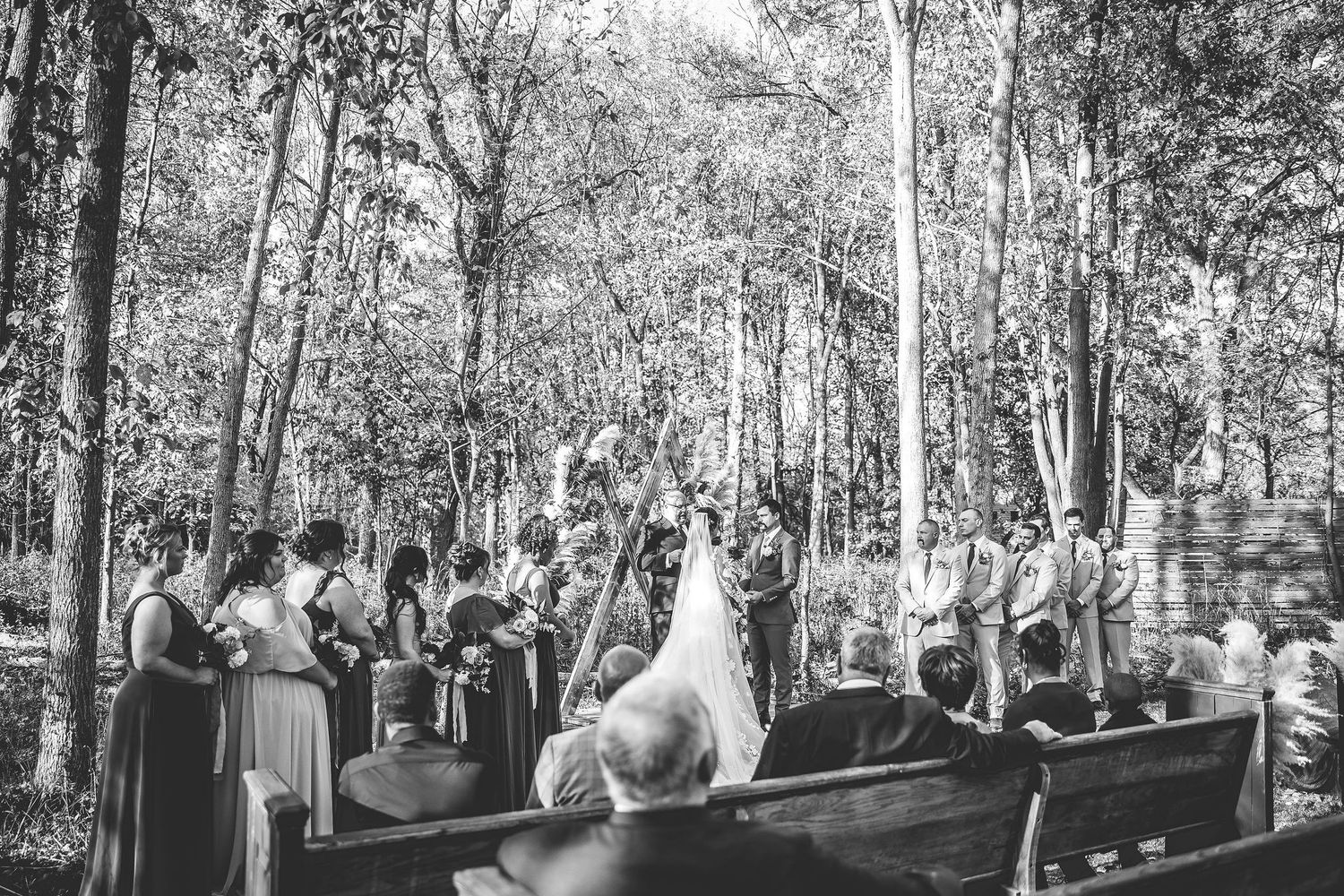 Black and white photograph of wedding ceremony in wooded setting with guests seated on benches.