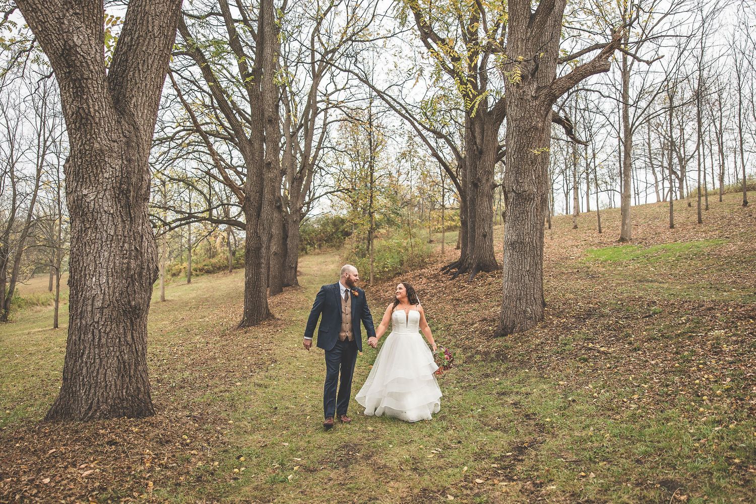 Couple walks hand in hand through a scenic wooded area during fall wedding photography session.