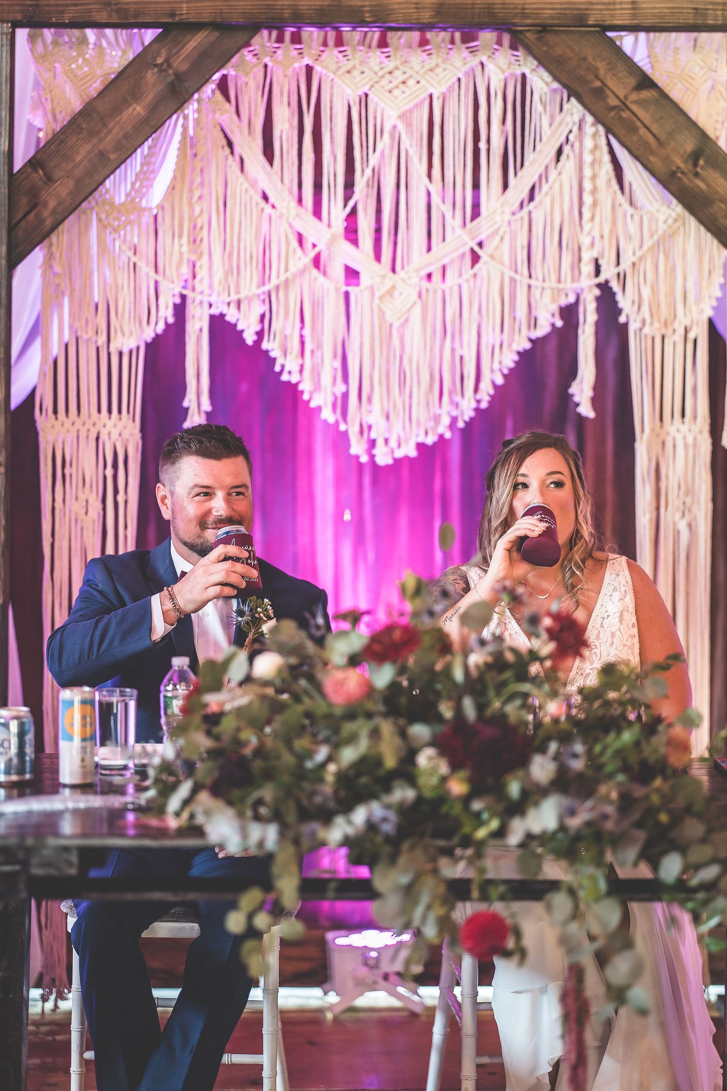 Wedding party delivers toasts at head table decorated with floral arrangements and macrame backdrop.