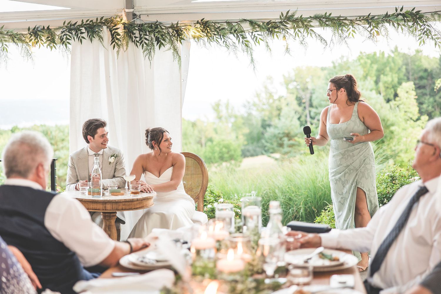 Wedding reception with guests listening to toasts under a decorated tent.