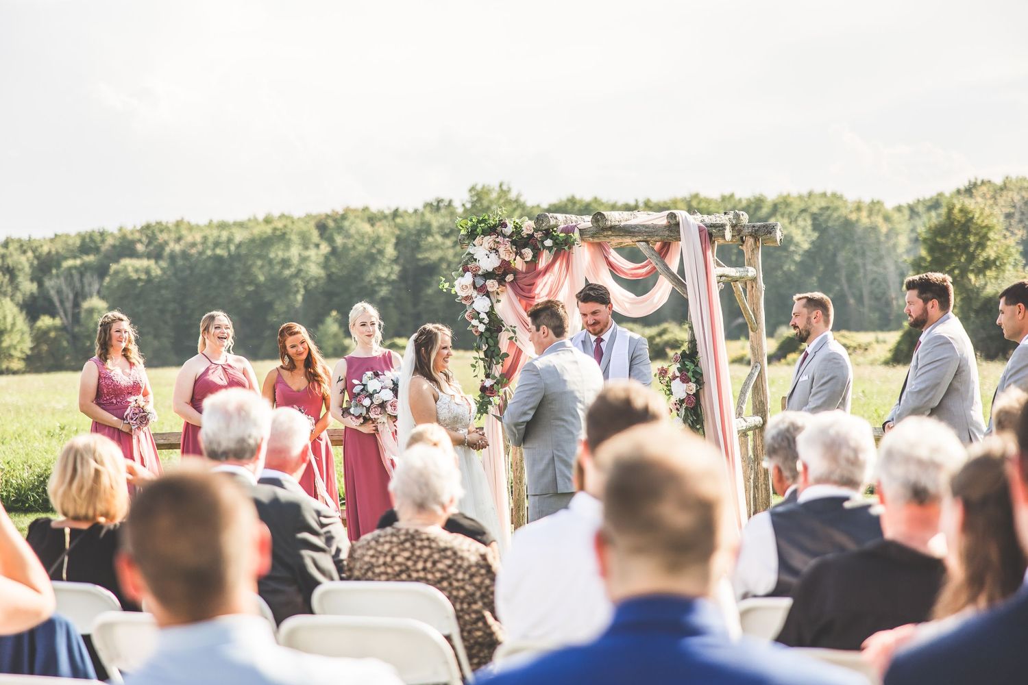 Wedding guests watching ceremony from white chairs on grass with trees in background.