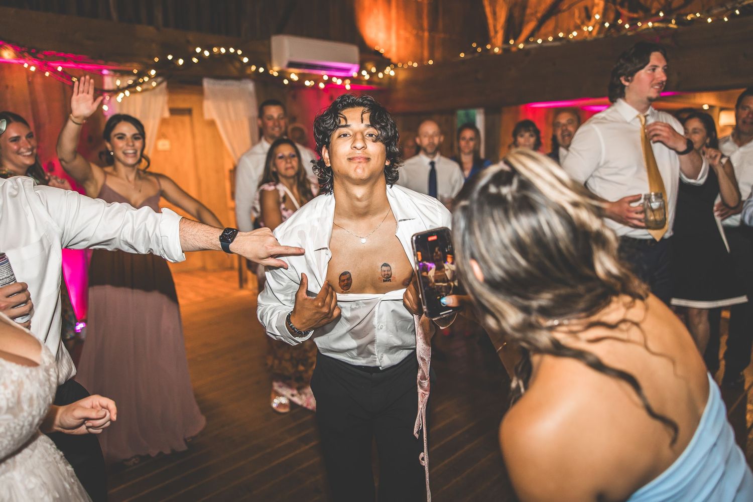 Wedding reception dance party with colorful lighting and string lights in a rustic indoor setting.