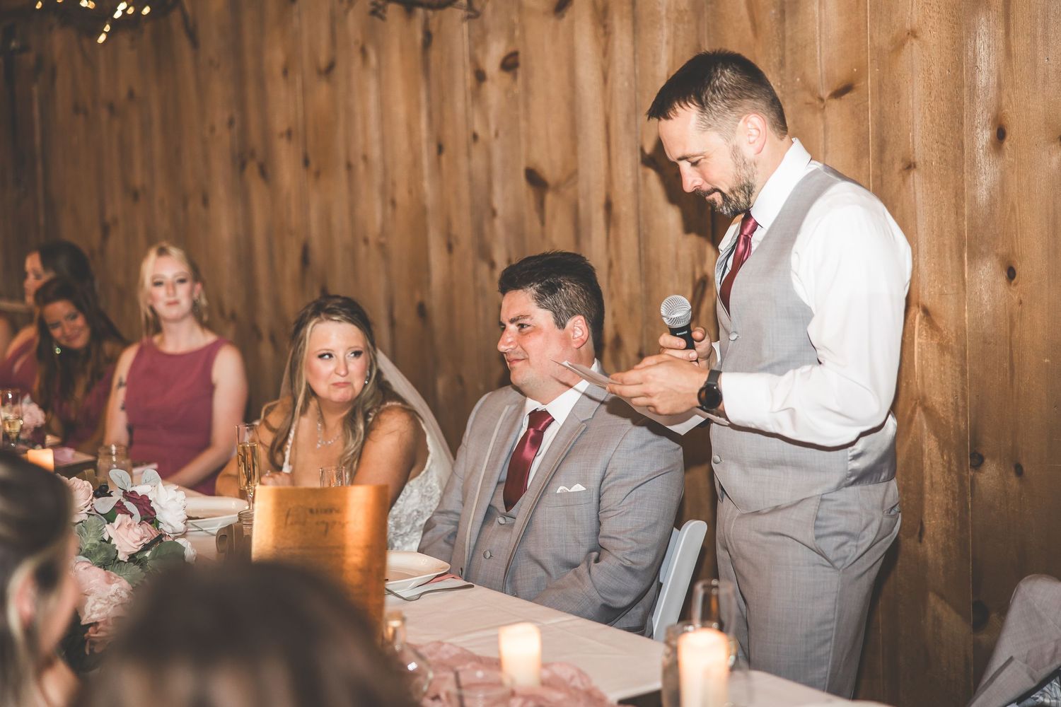 Wedding reception toast being given in a rustic wooden barn venue with string lights and table decorations.