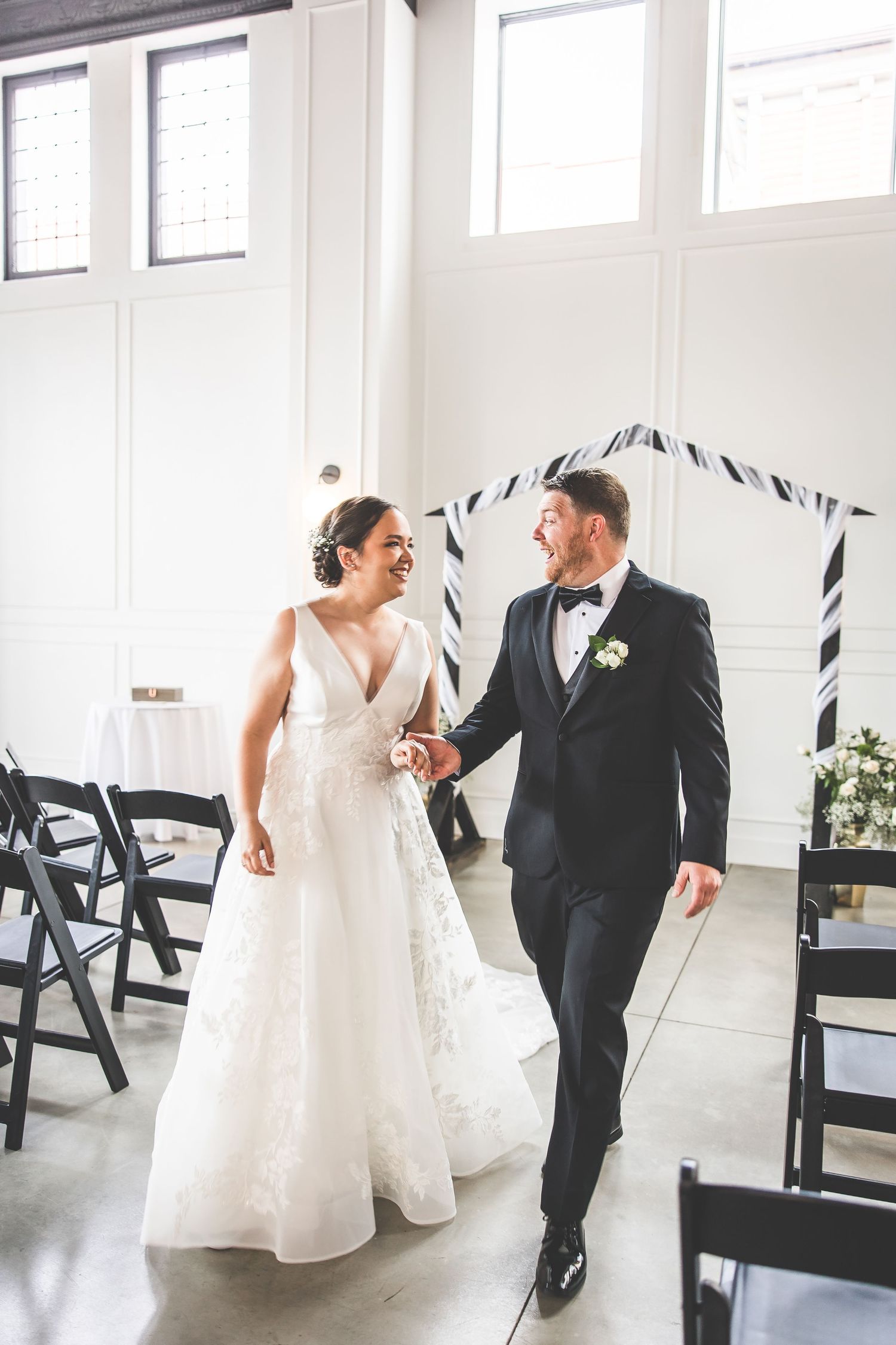 Couple sharing playful dance moves in a modern white venue with black folding chairs.