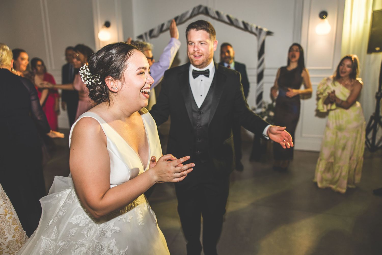 Elegant wedding reception with guests dancing in a white-walled venue with pendant lighting.