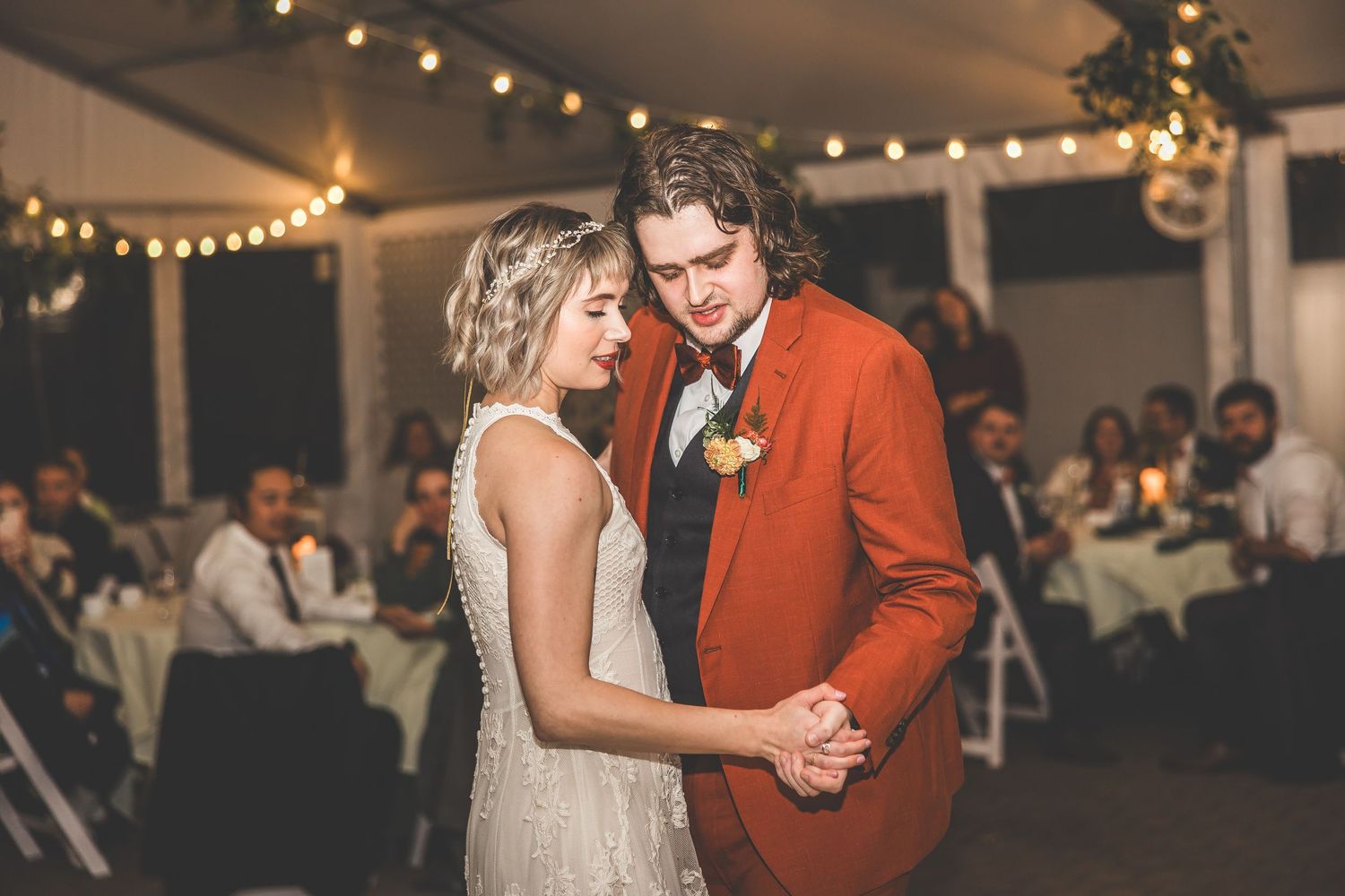 Wedding guests dance and celebrate on a wooden floor under string lights and tent canopy.