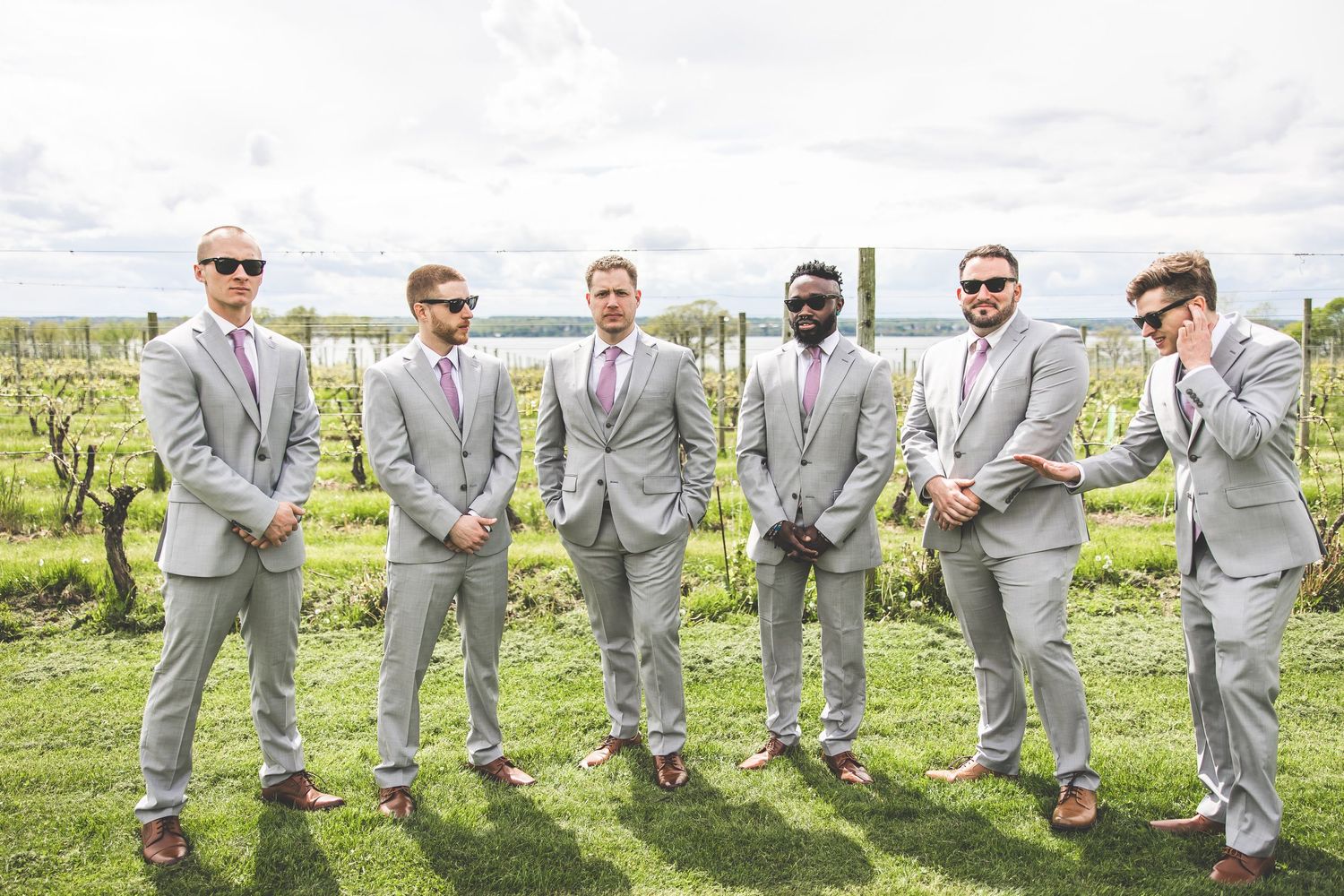 Groomsmen in light gray suits strike playful poses in a row against a vineyard backdrop.
