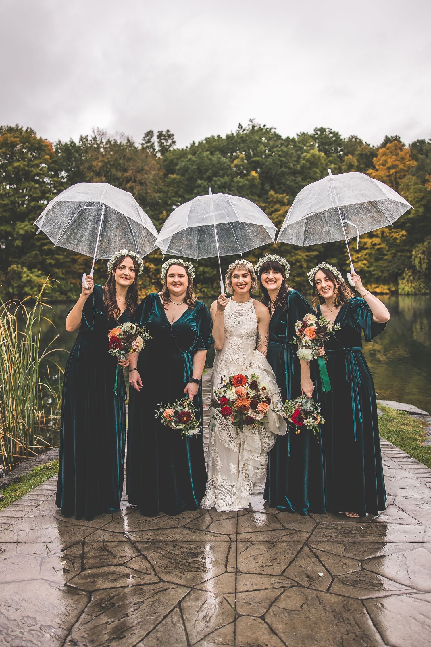 Wedding party with clear umbrellas poses on stone patio by lake in velvet dresses.