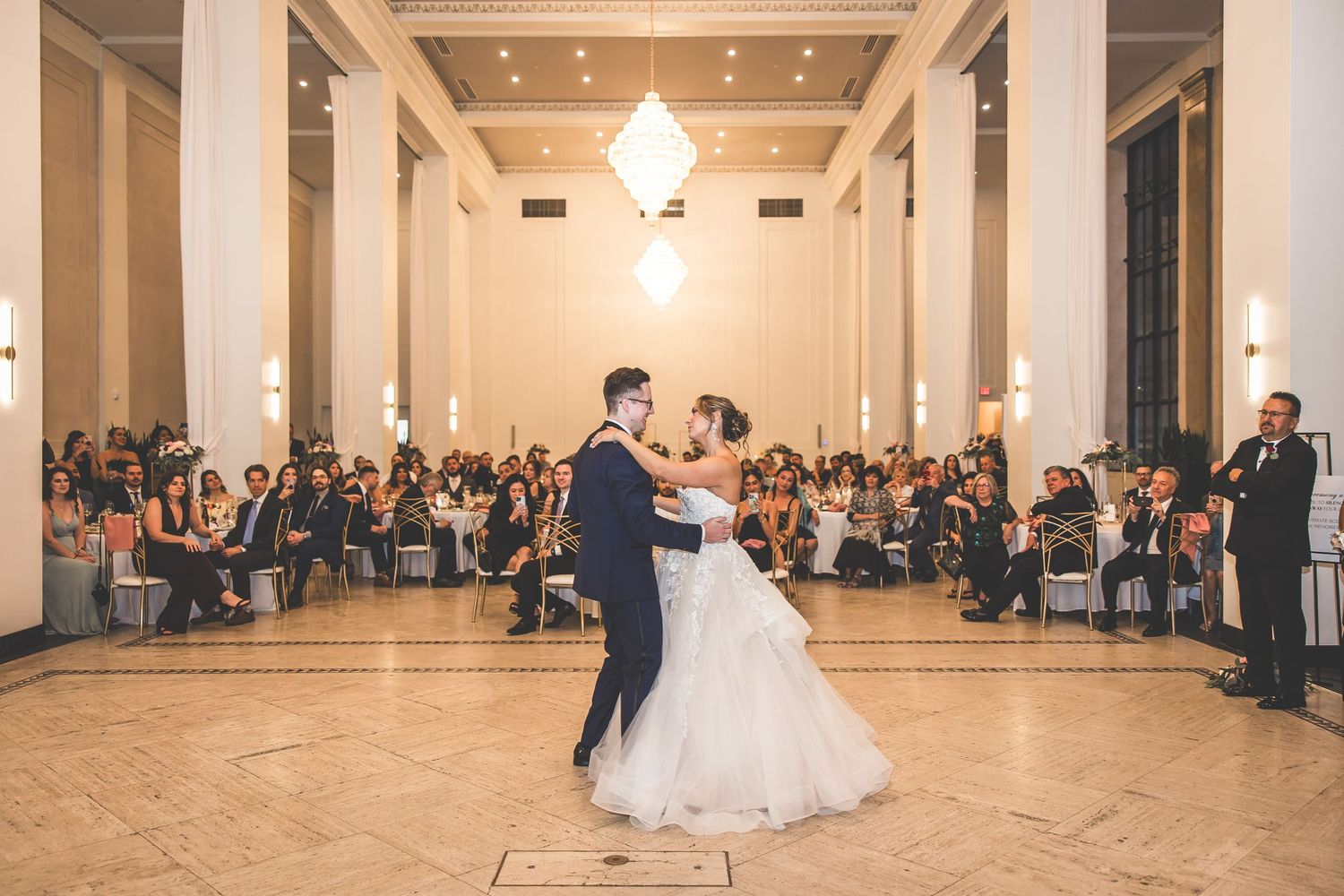 Wedding couple dances in spacious ballroom while guests observe from the sides.