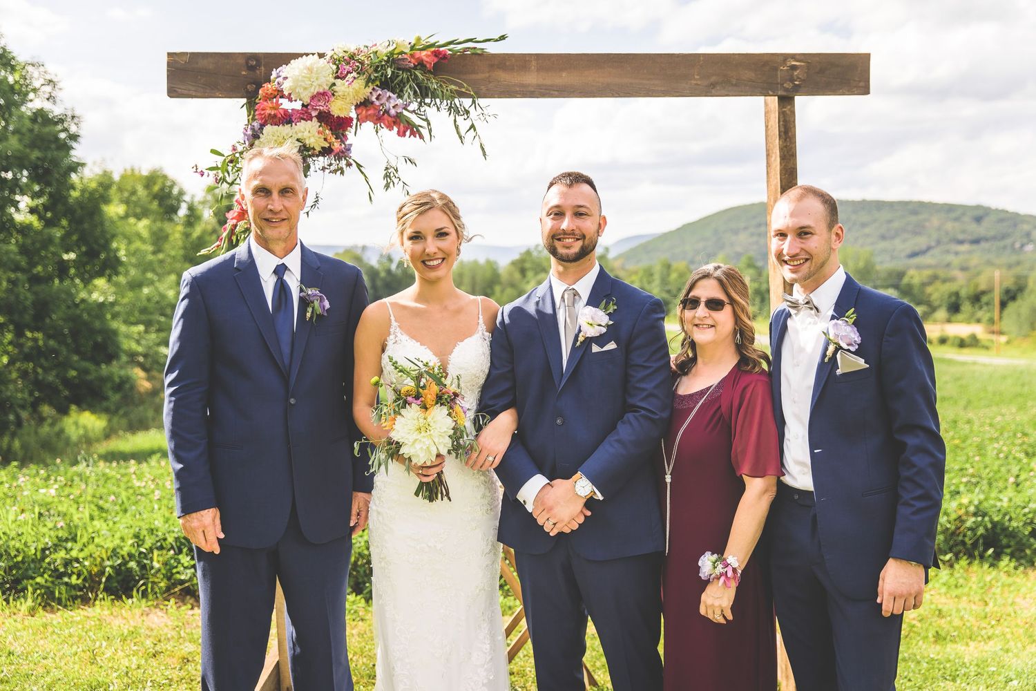 Wedding party group photo under wooden arch with mountains in background.