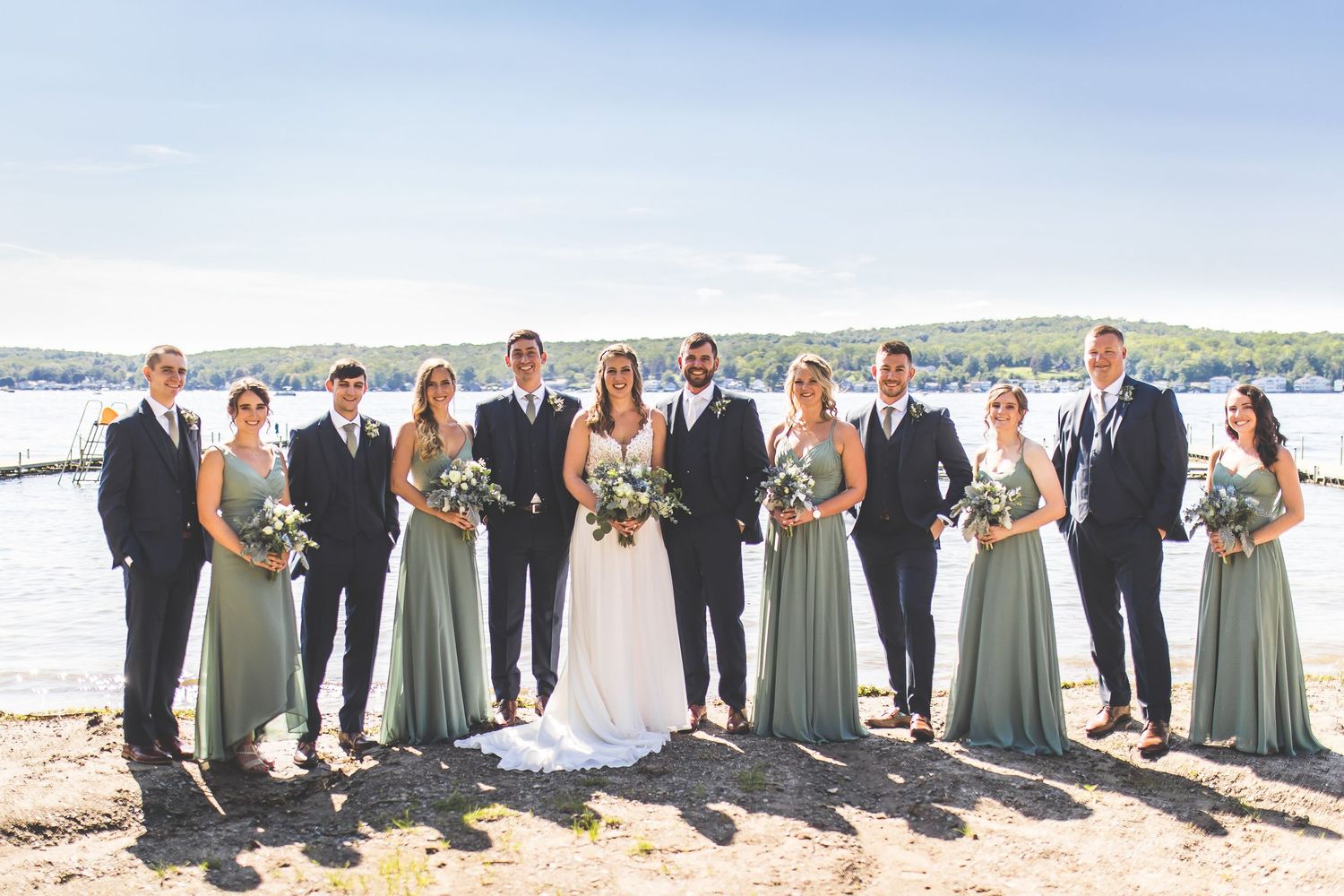 Wedding party in sage green dresses and dark suits poses together by a waterfront setting.