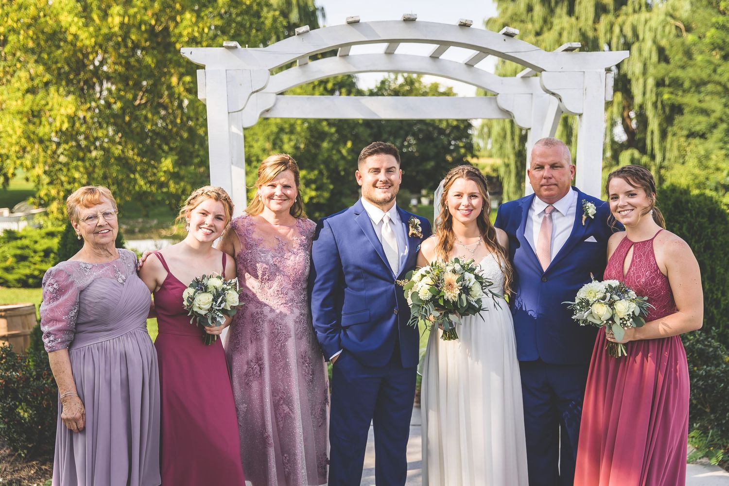 Wedding party members in burgundy dresses and navy suits pose beneath a decorative white arbor.