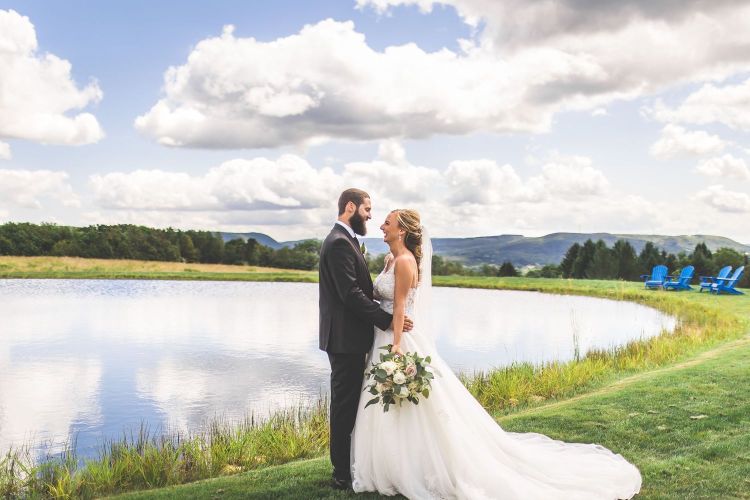 Wedding couple poses by a serene lake with mountains and clouds in the background.