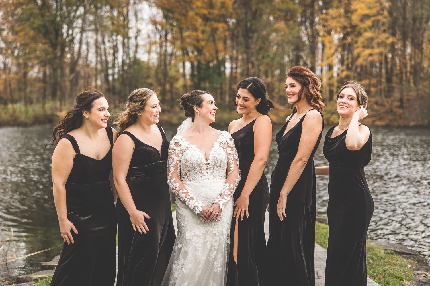 A bridal party in black dresses poses with the bride in a white lace gown by a lake during autumn.