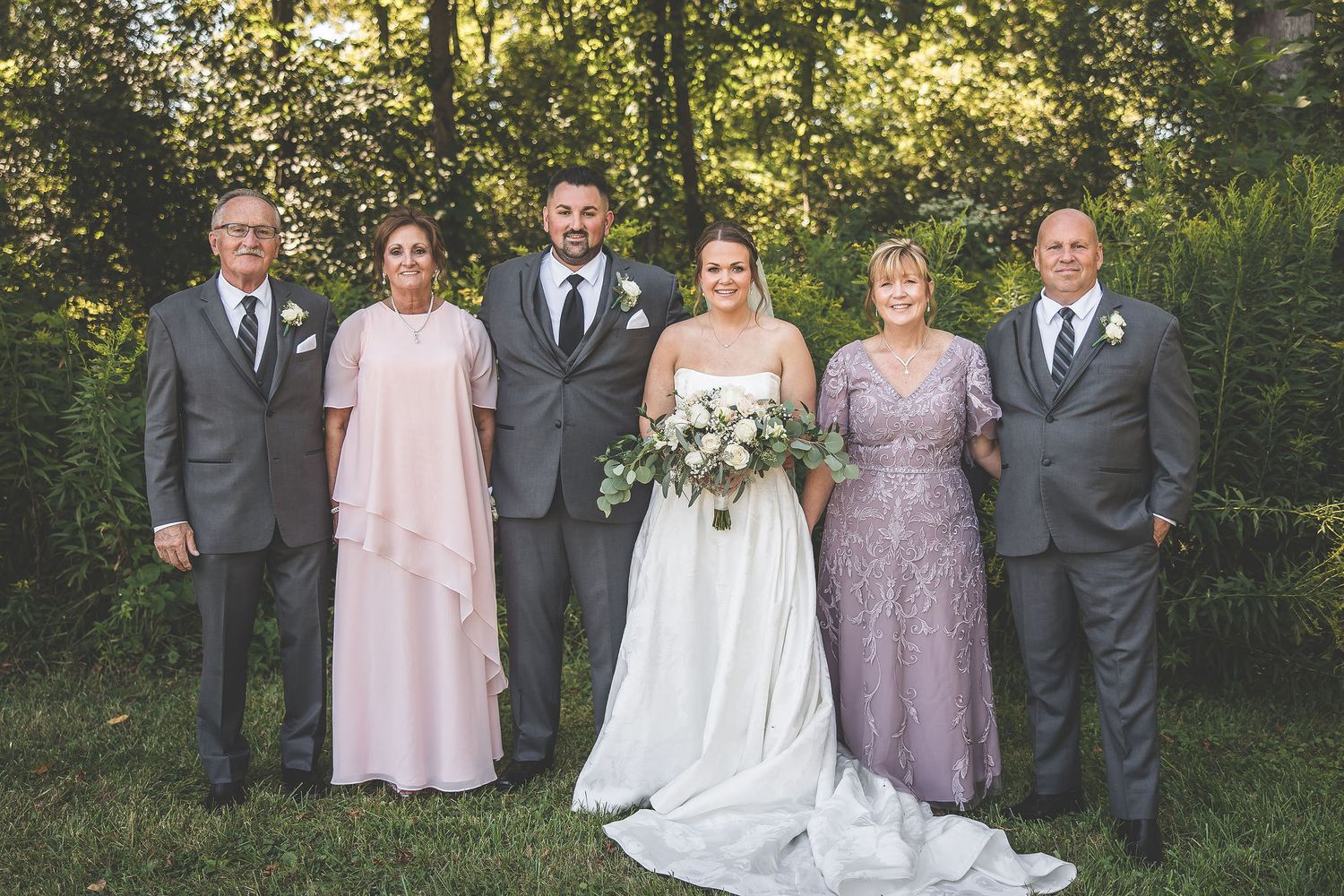 Wedding party in gray suits and pastel dresses poses formally against a natural outdoor backdrop.