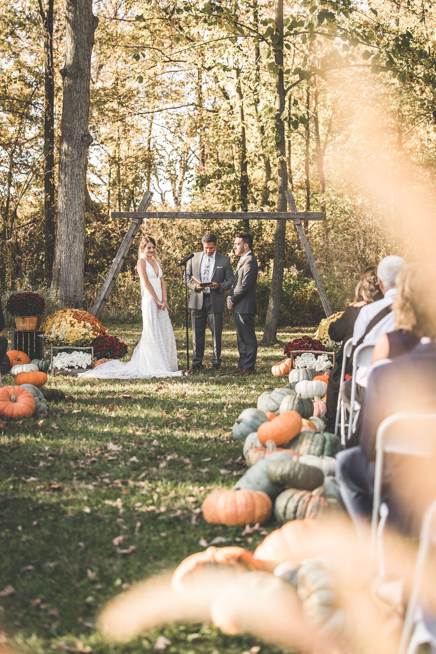 Outdoor autumn wedding ceremony with rustic wooden arch and pumpkin decorations in a wooded setting.