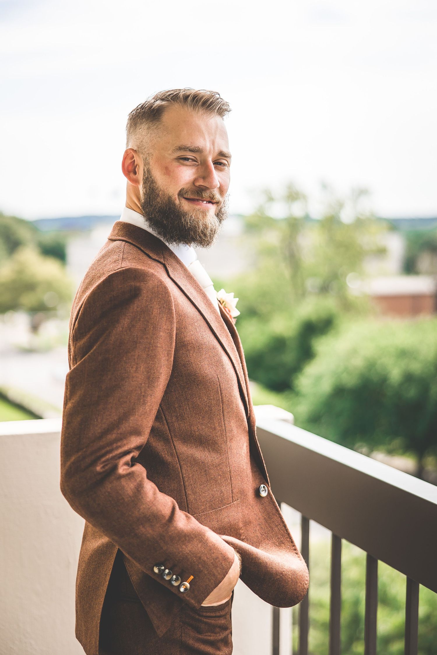 Person wearing brown tweed jacket poses on outdoor terrace with greenery in background.