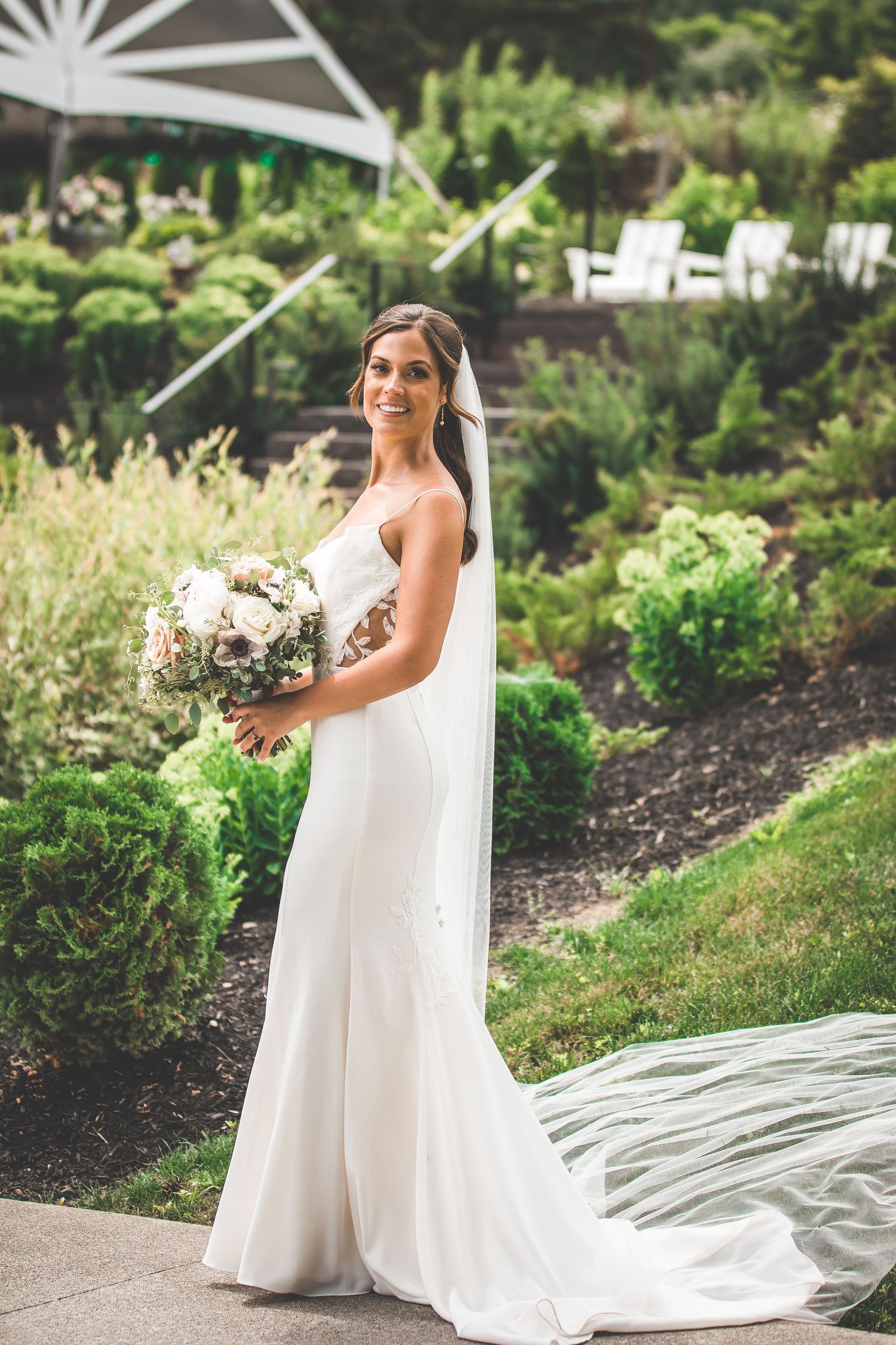 Bride in flowing white gown with long train poses in a garden setting with landscaped flower beds.