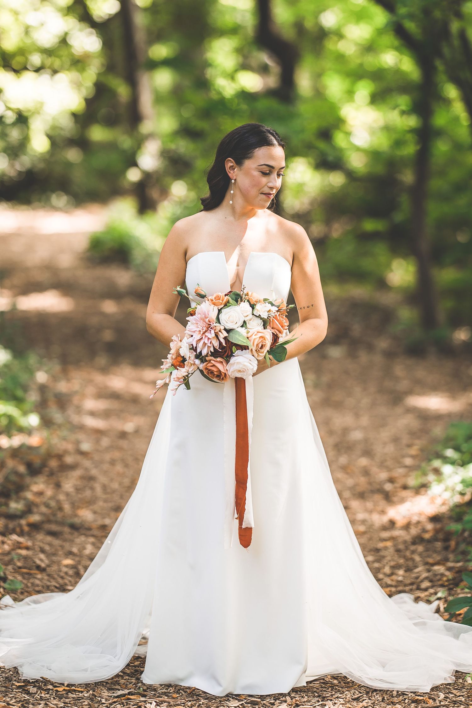 Bride in elegant white gown holds bouquet in sunlit forest setting.