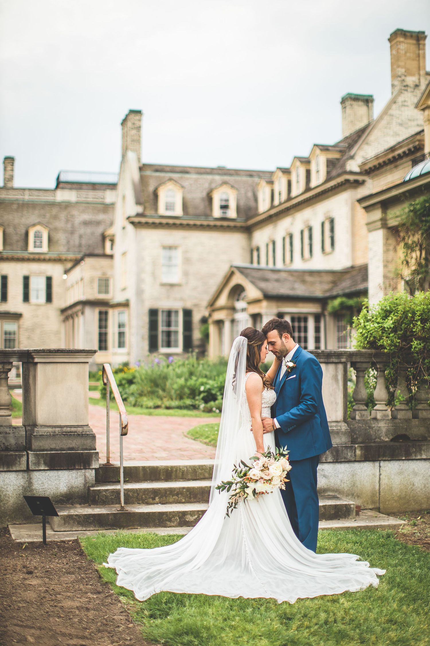 A wedding couple shares a romantic moment on the steps of a grand historic mansion.
