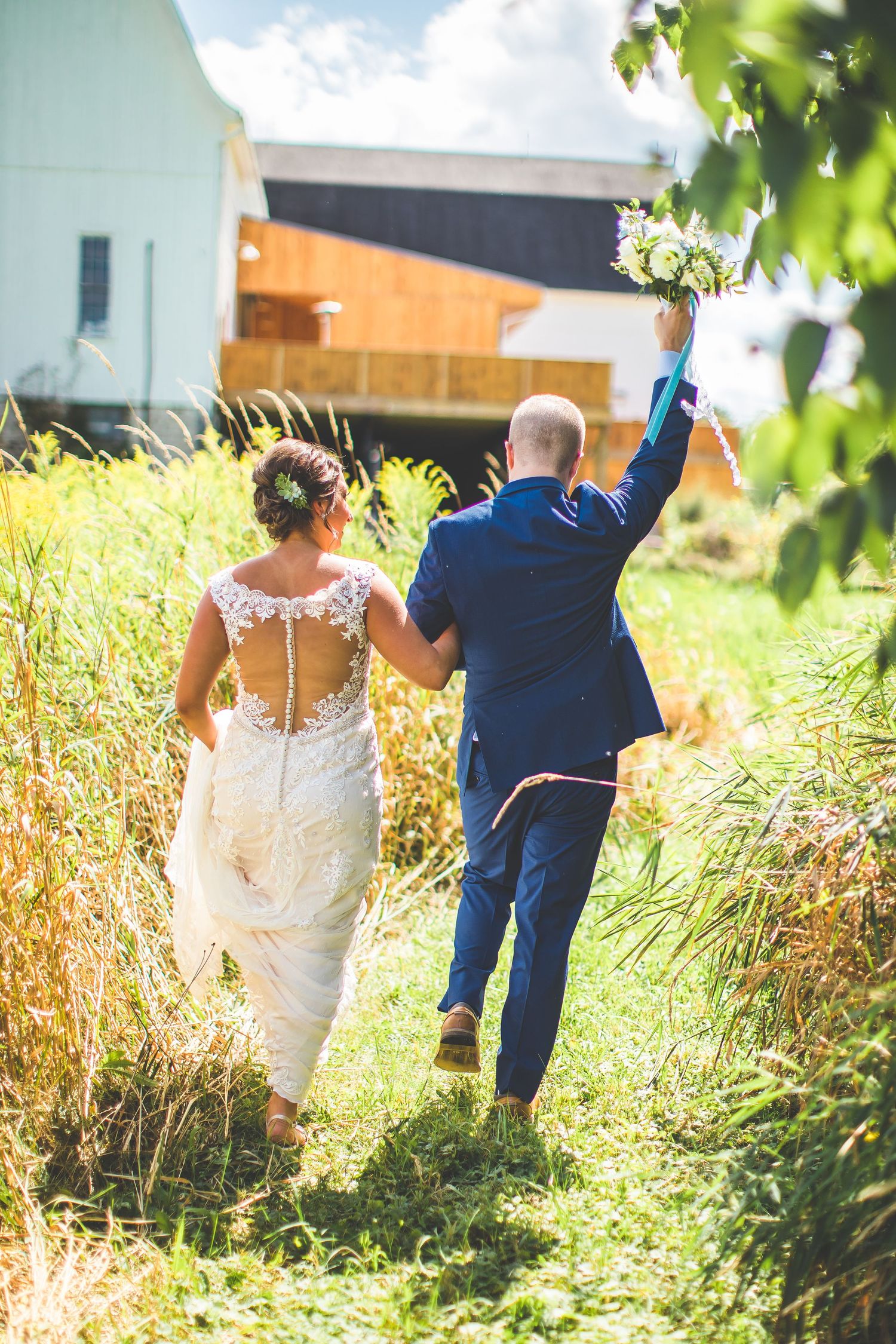 Couple walking through tall grass in wedding attire on a sunny day.