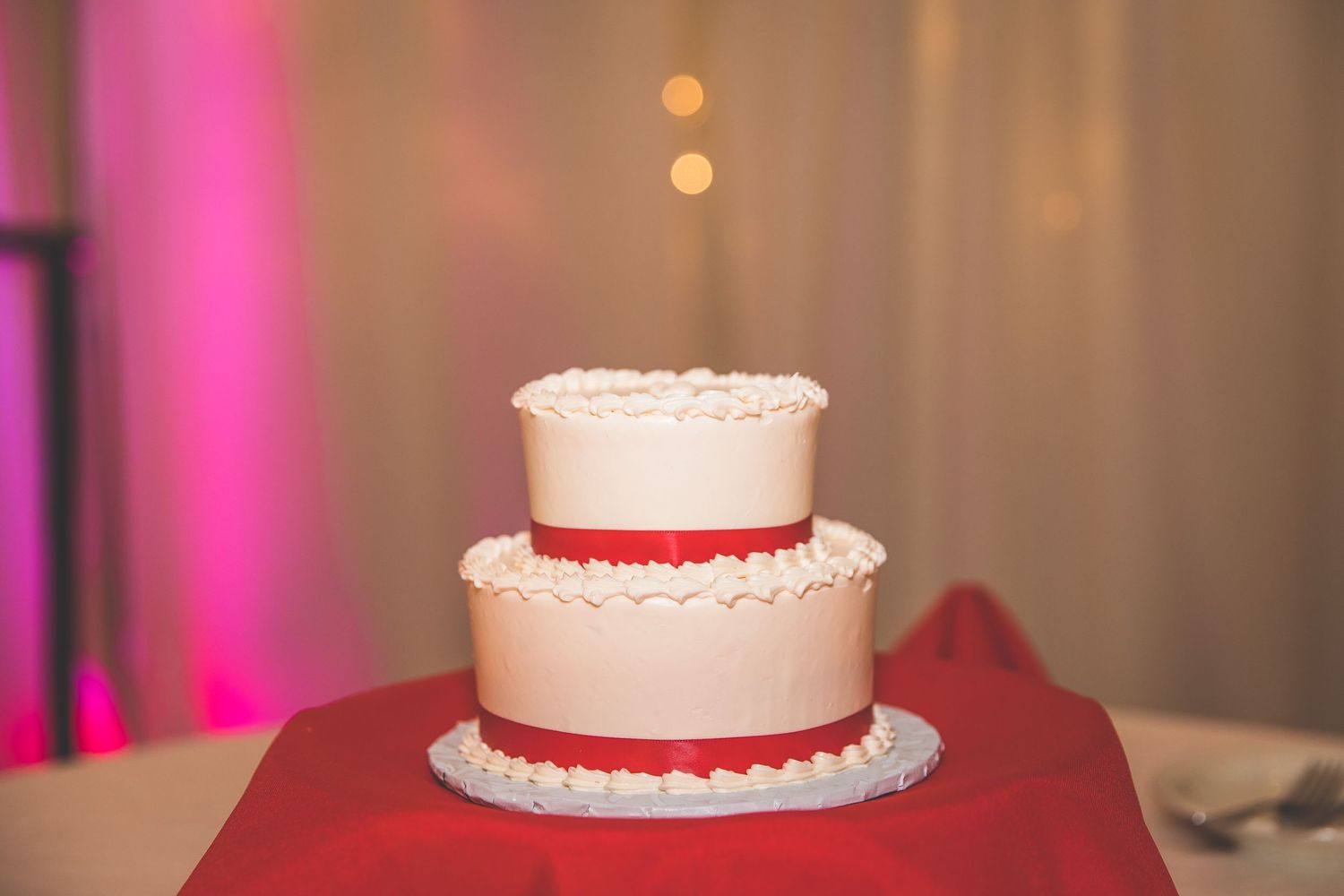 White wedding cake with delicate lace details displayed on a red tablecloth.