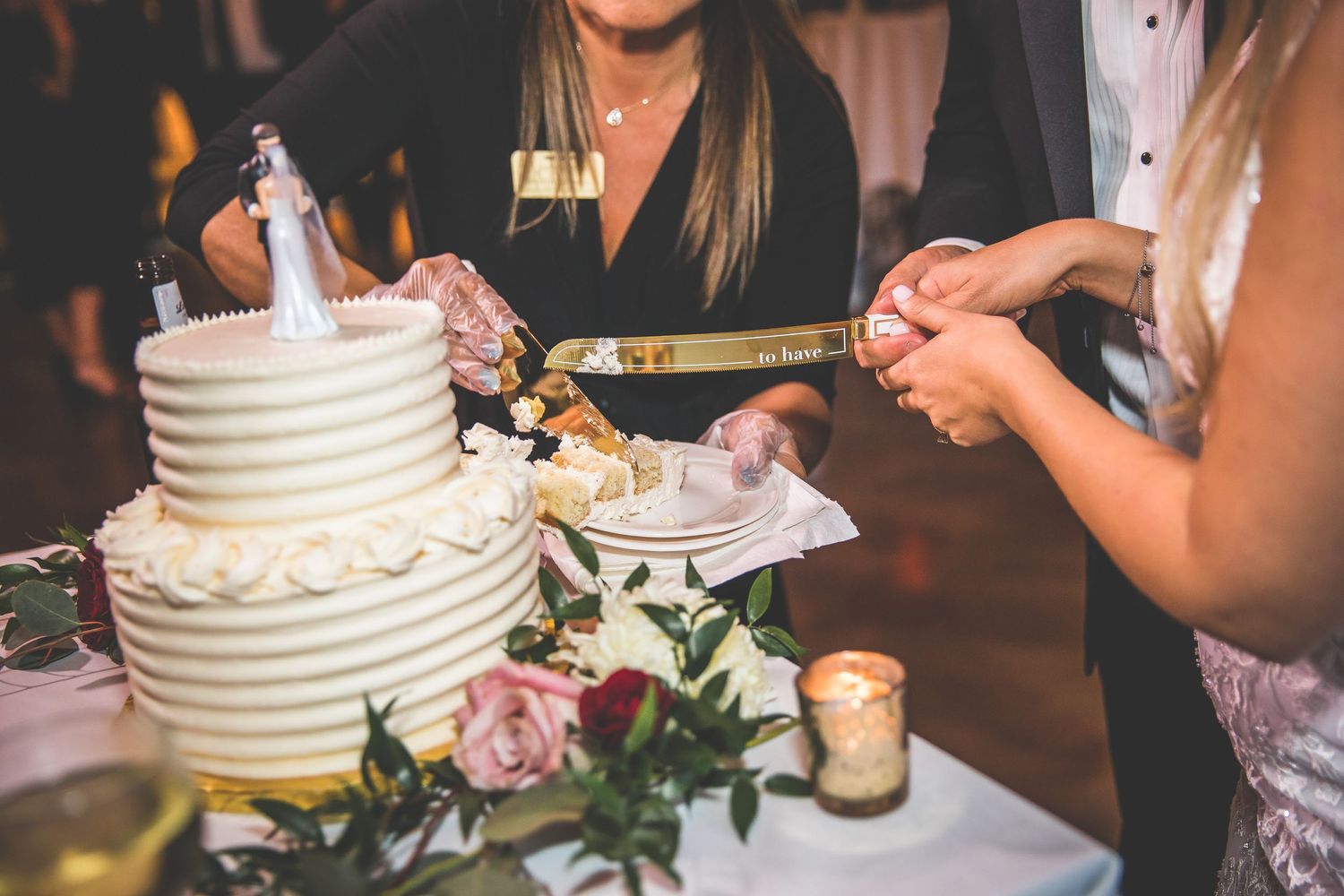 Close-up of hands cutting a white tiered wedding cake decorated with roses.