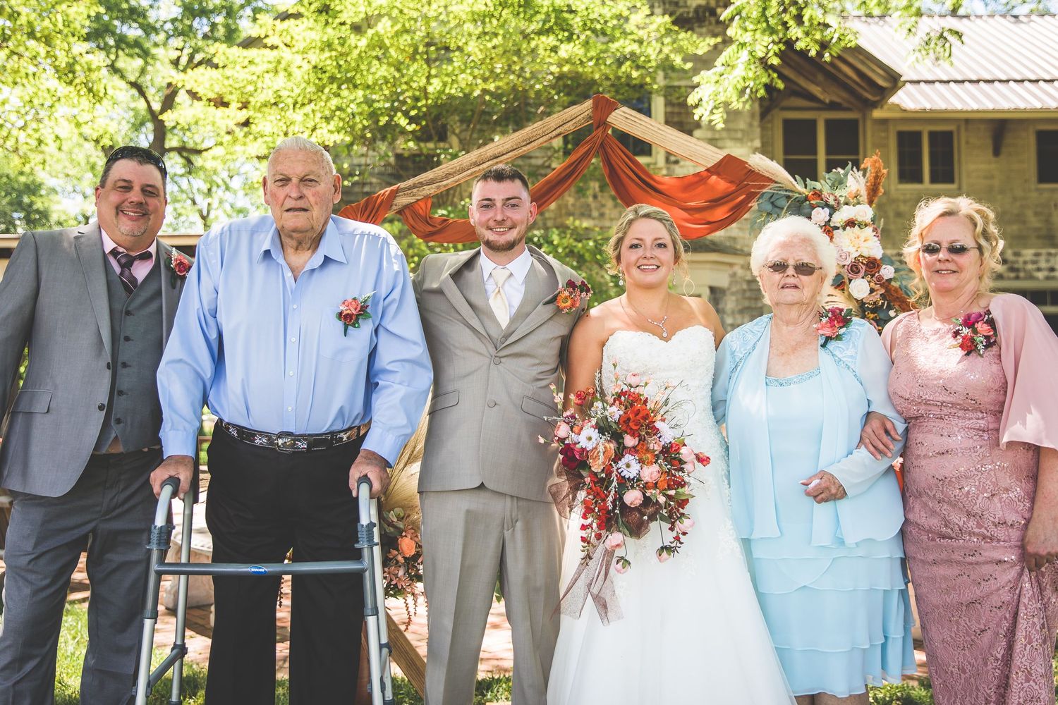 Wedding party poses outdoors with decorative arch and floral arrangements.