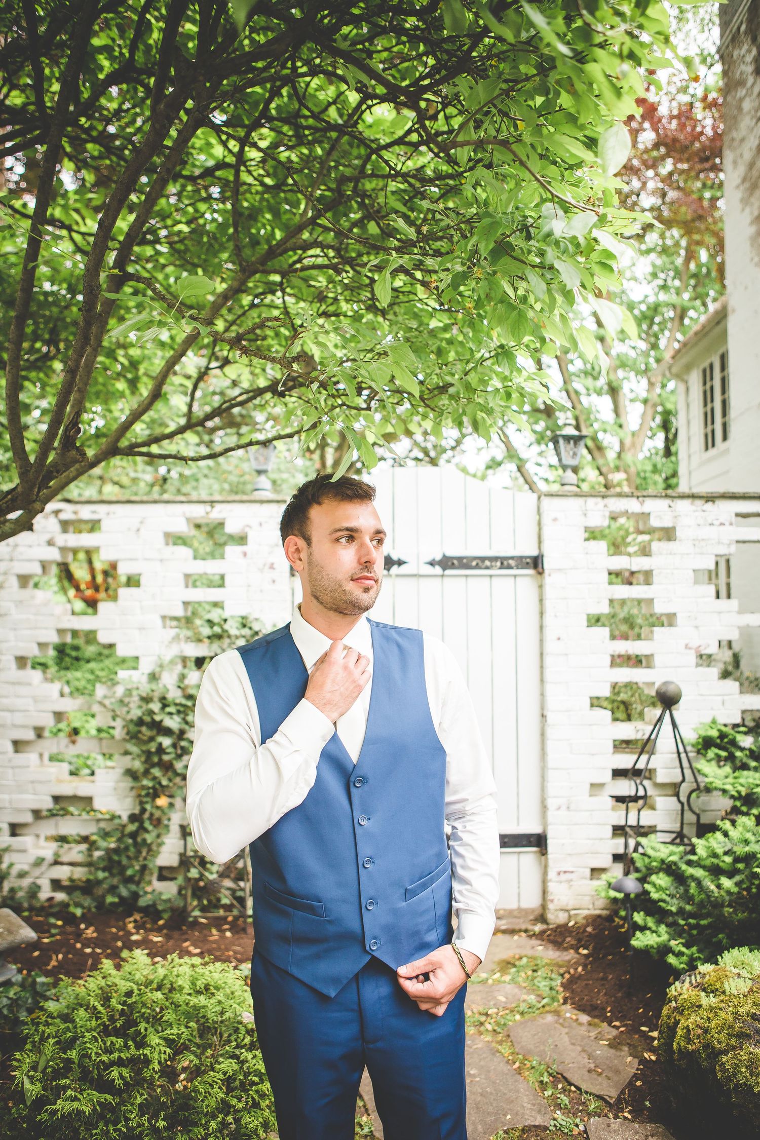 Groom in blue vest standing among greenery and white brick walls.