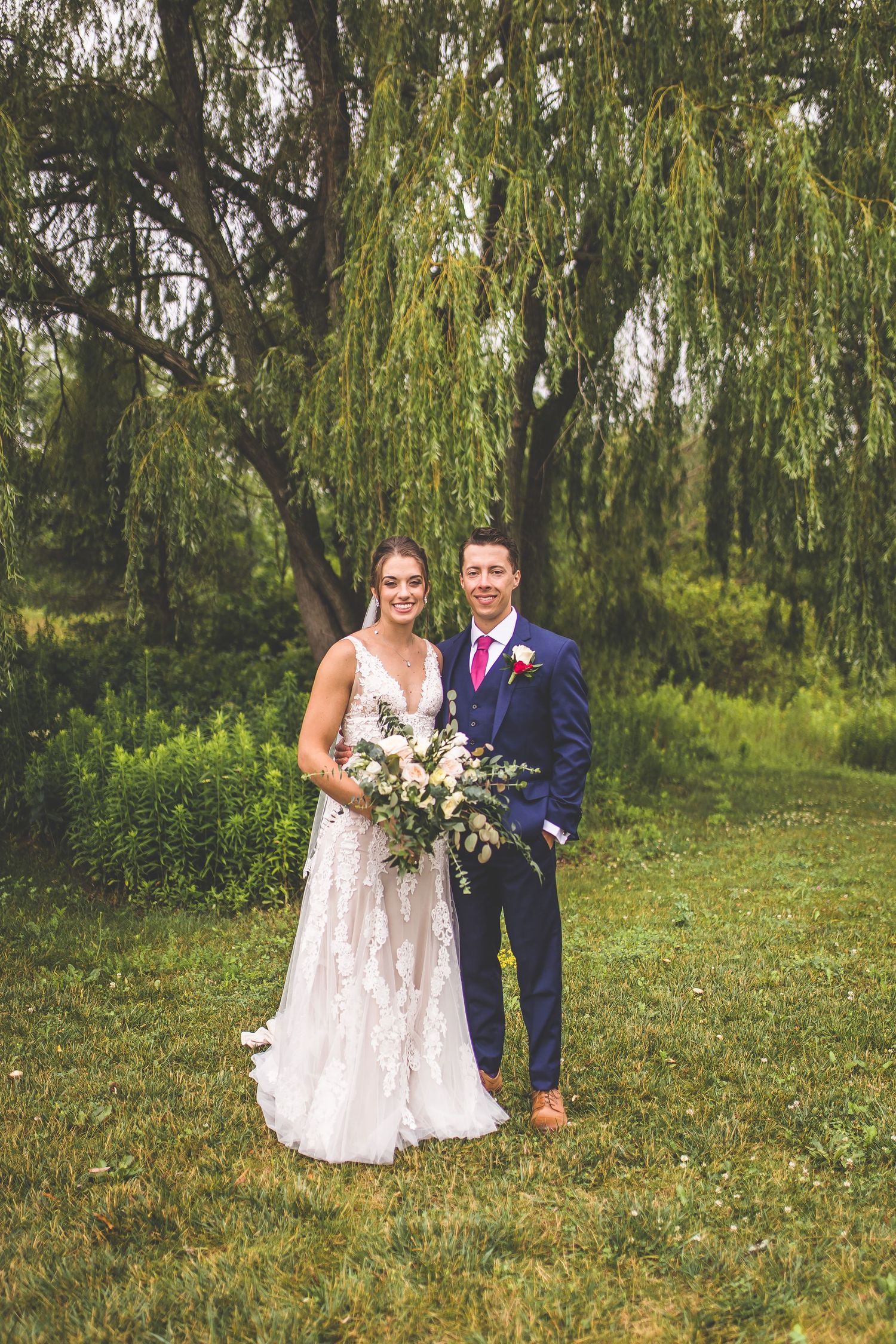 A wedding couple poses together under a beautiful weeping willow tree in a garden setting.