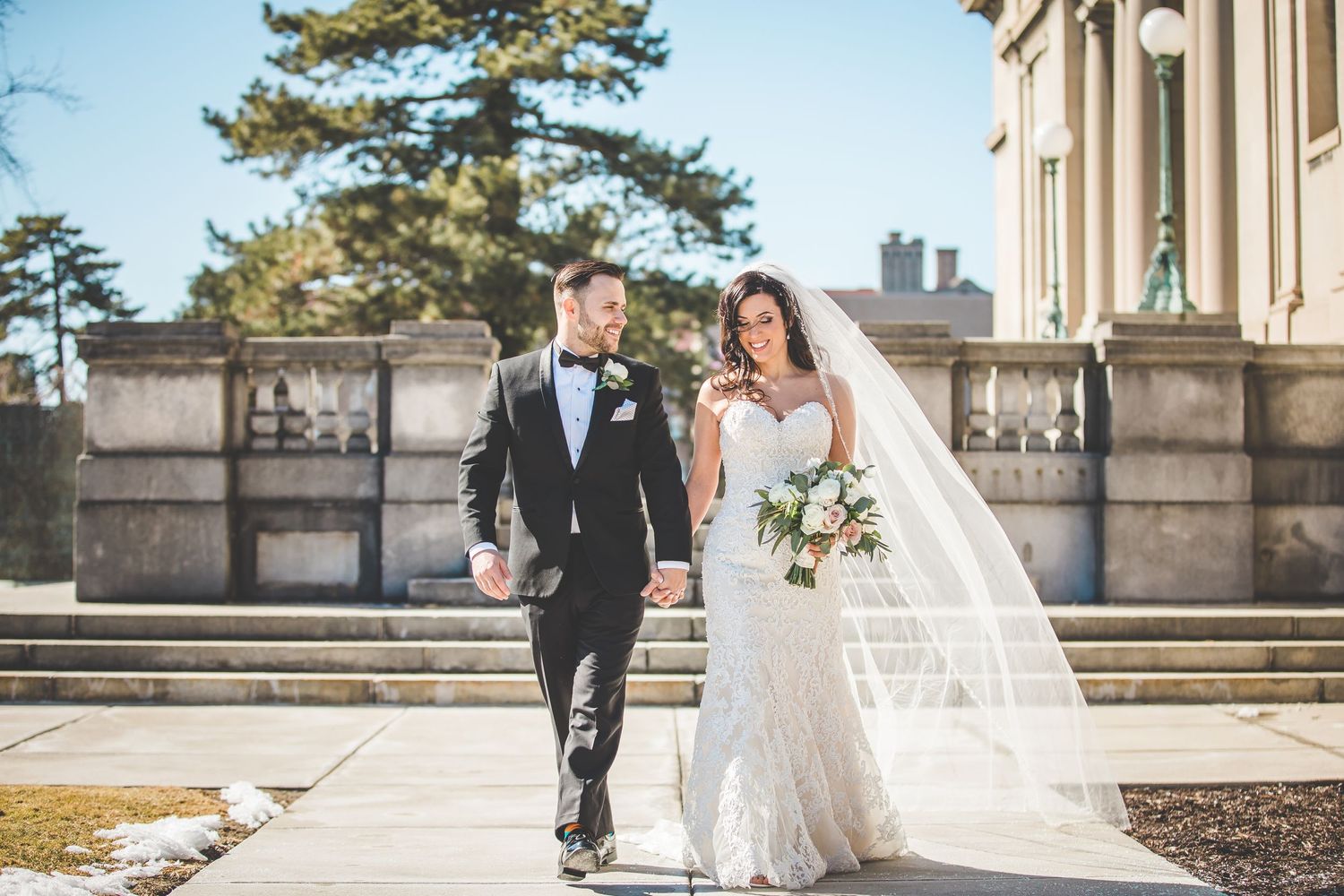 A couple walks together on stone steps outside a grand building on their wedding day.