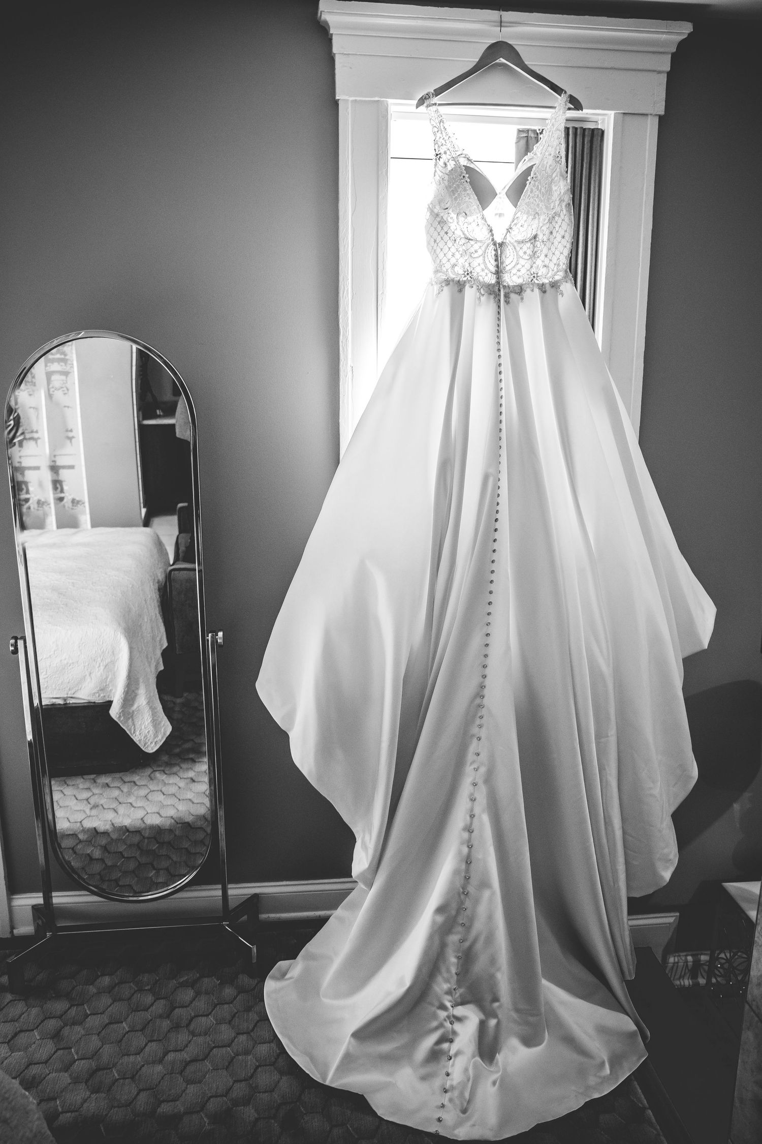 A white wedding dress with buttons hangs elegantly against a gray wall in natural window light.