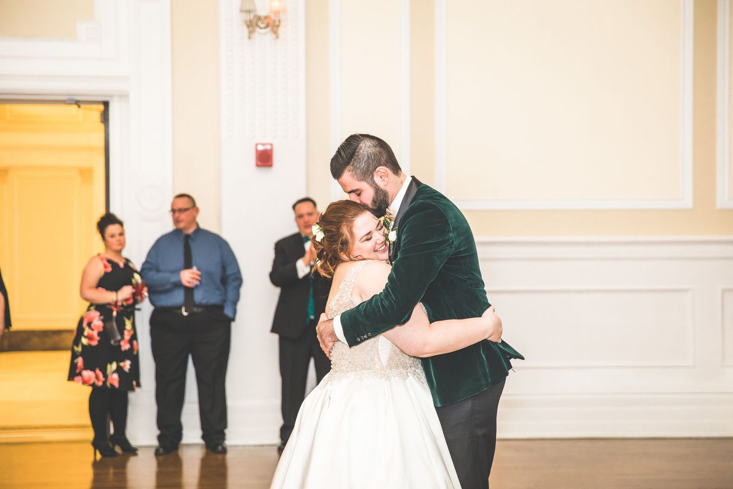 Wedding party members dance and celebrate in an ornately decorated ballroom with cream-colored walls.