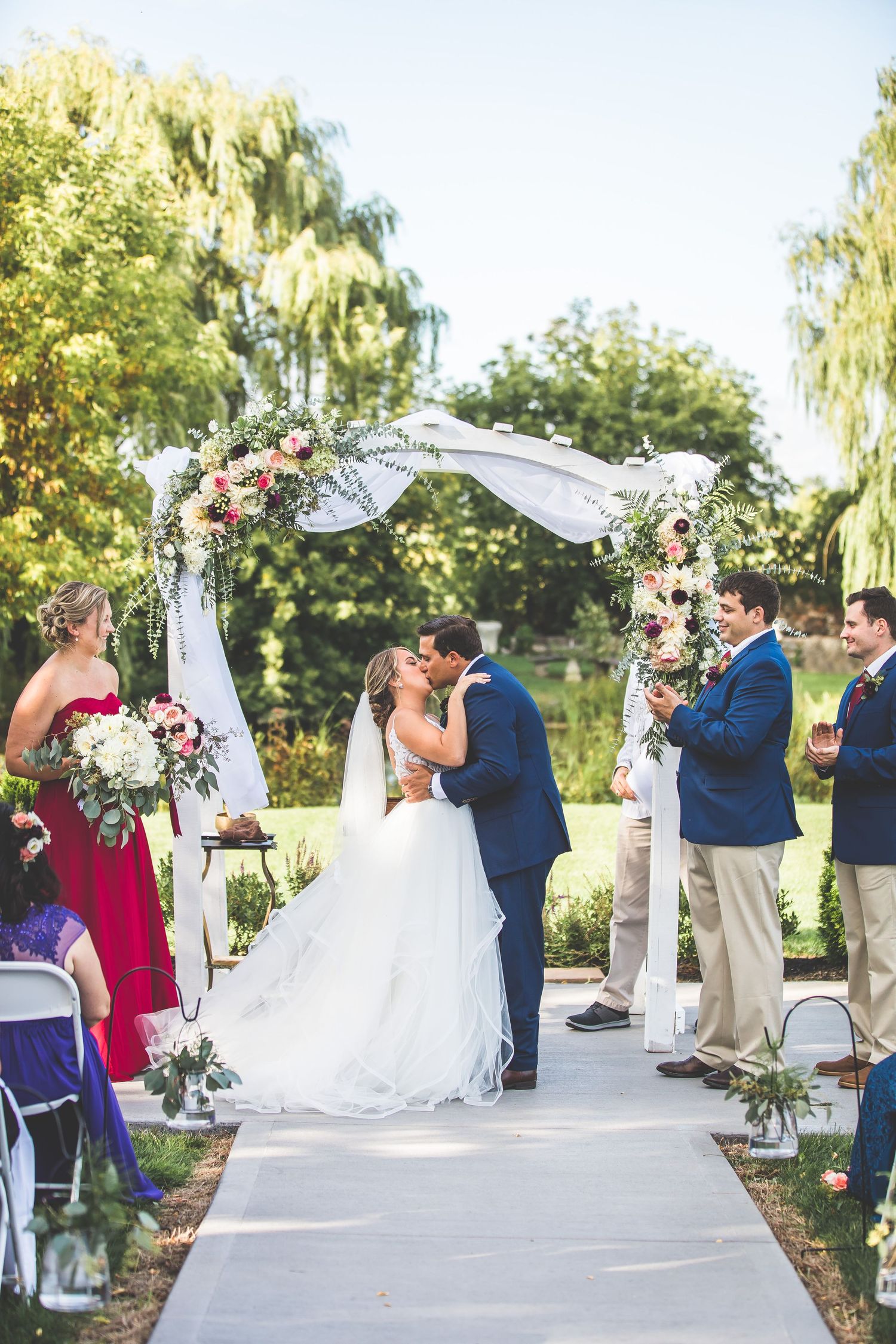 Outdoor garden wedding ceremony under a white draped arch with guests seated on white chairs.
