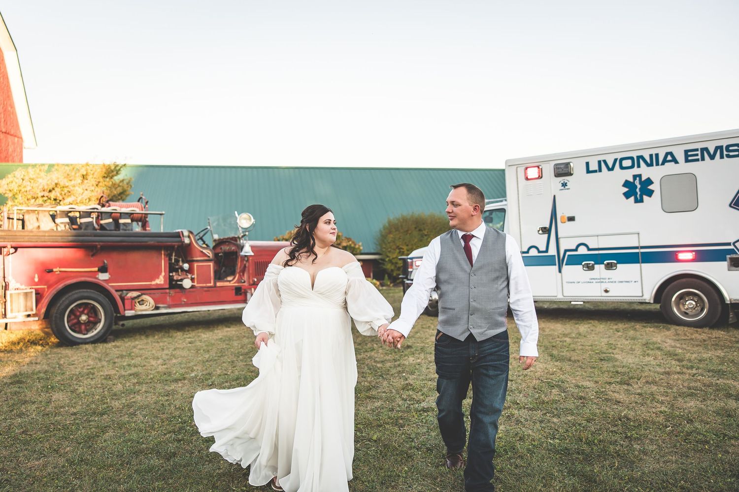 Outdoor wedding celebration on grassy field with emergency vehicles in background at sunset.