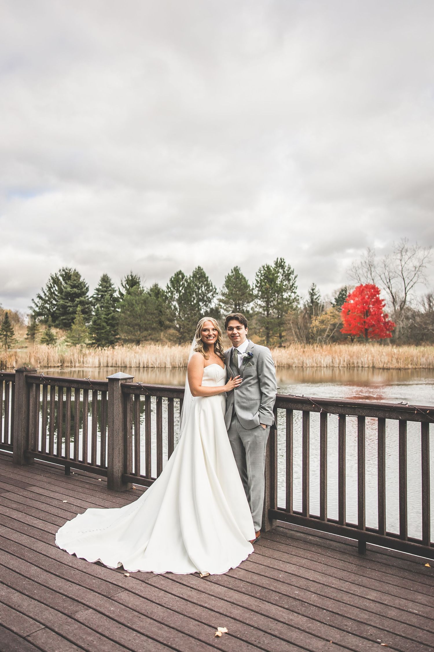 Newlyweds embrace on wooden deck with fall foliage and lake in background.