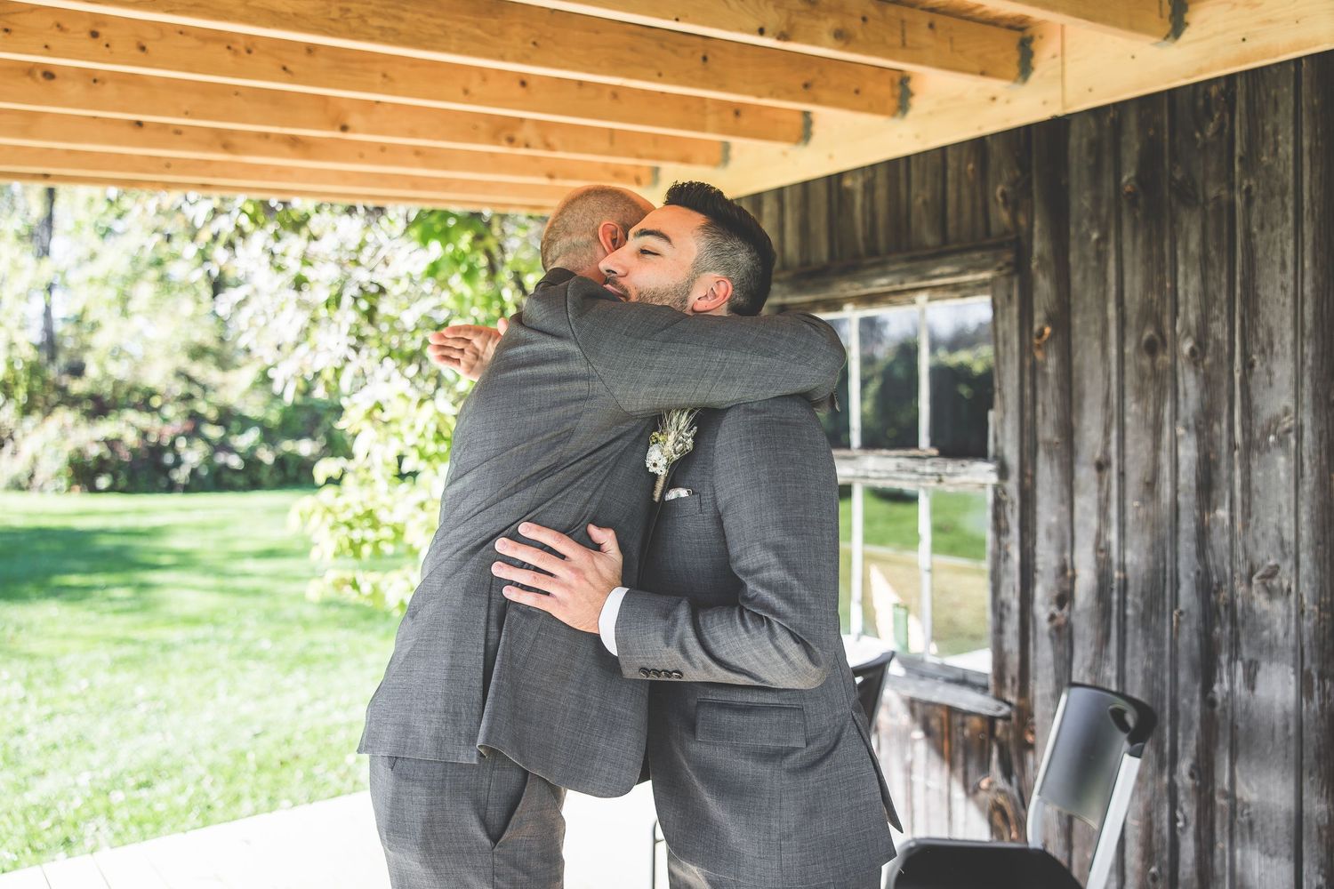 Two people in grey suits share warm embrace on covered porch during wedding day.