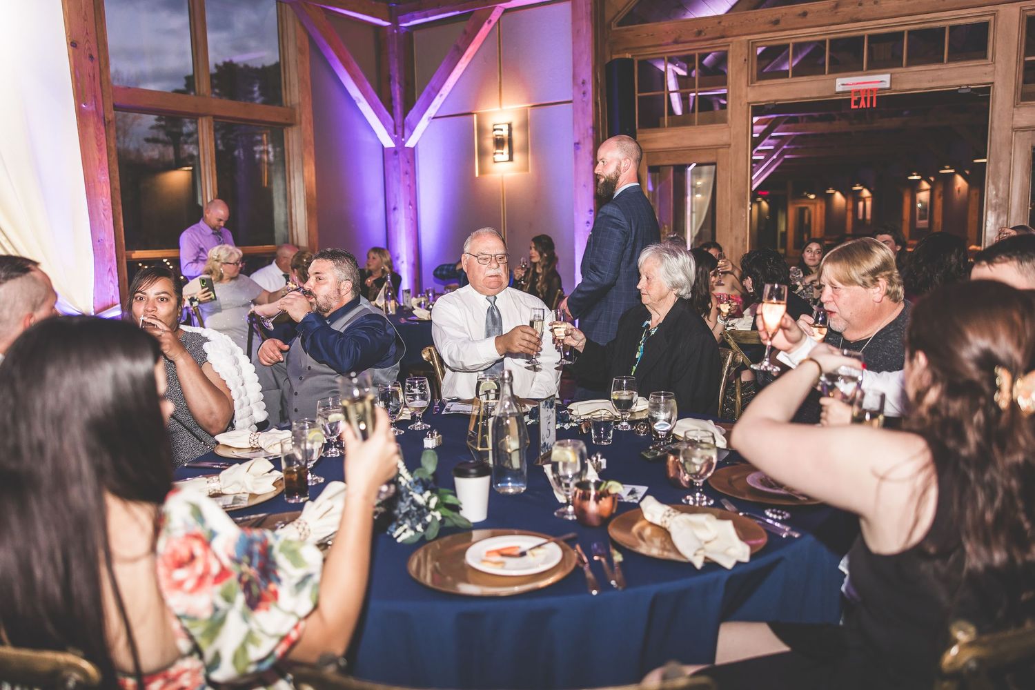 Wedding reception guests seated at round tables with navy linens and purple ambient lighting.