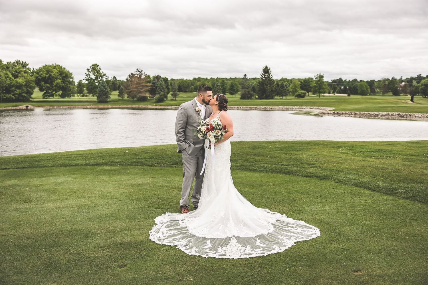 Bride's lace train spreads beautifully across green grass during outdoor wedding photos.