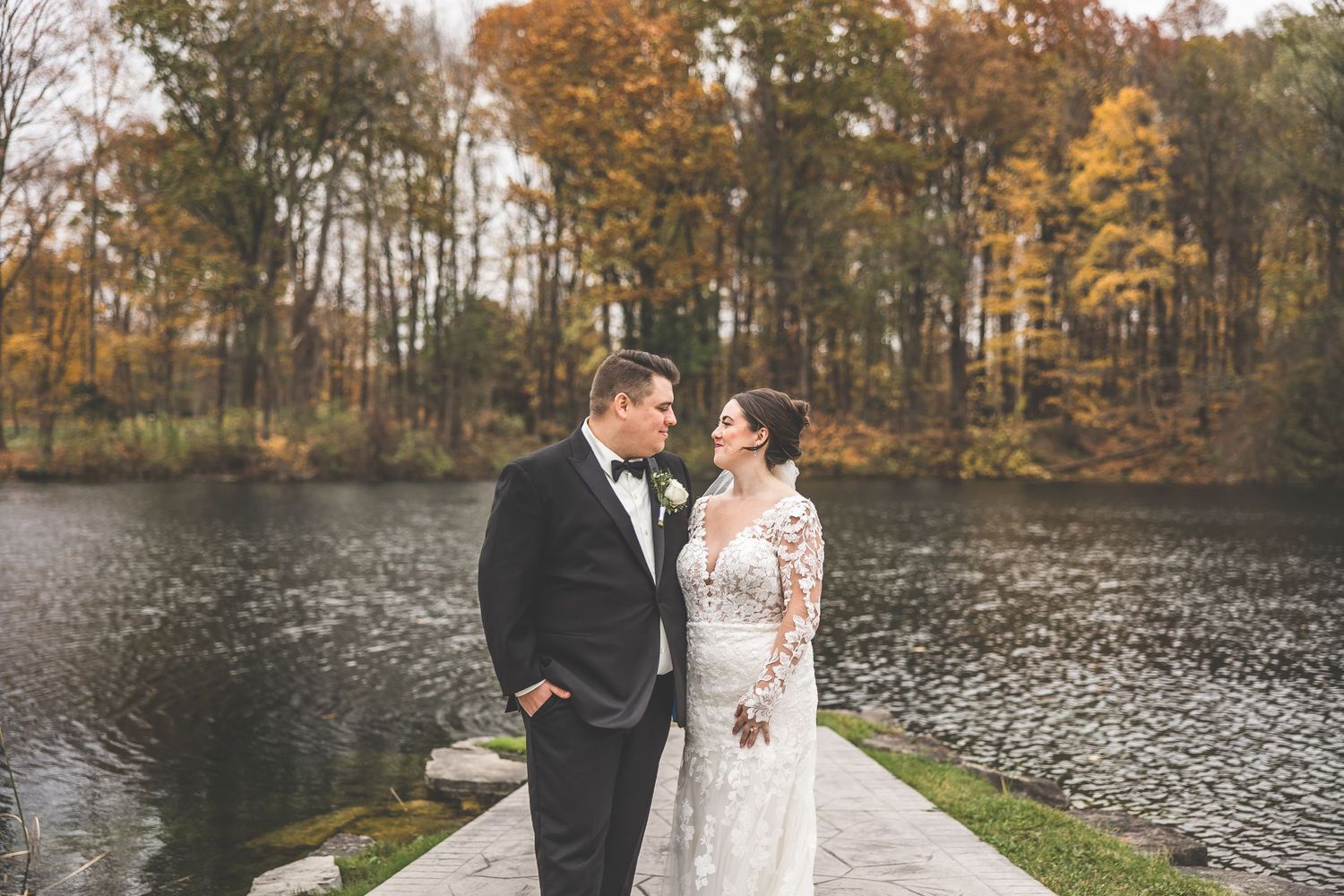 Romantic wedding portraits by lakeside with autumn trees and stone pathway.