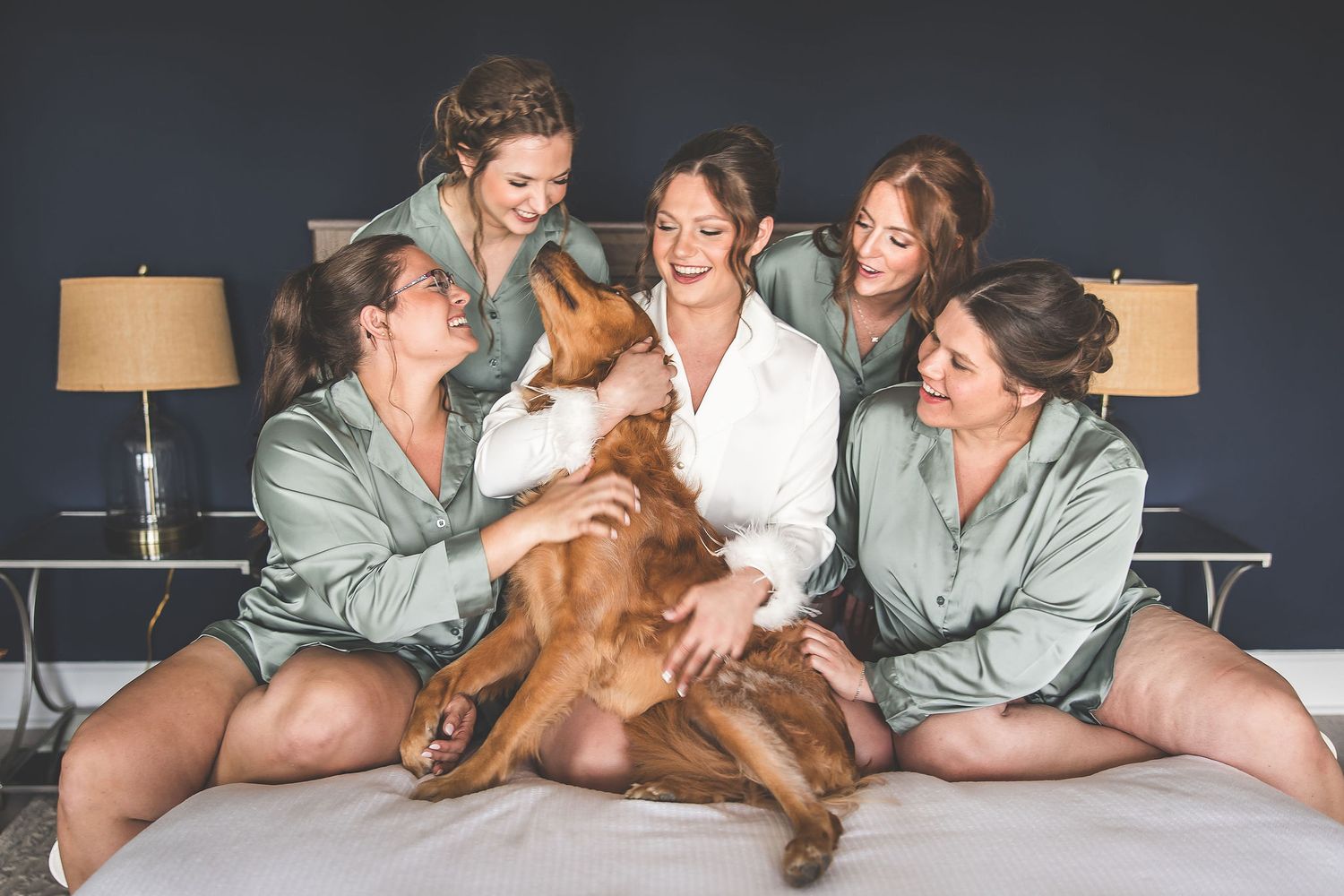 Friends in matching loungewear cuddle with a happy golden retriever on a bed with table lamps.