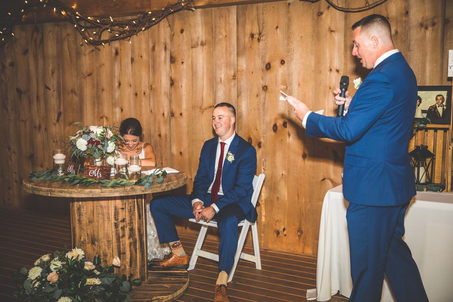 Wedding reception speech being delivered in a rustic wooden barn venue with floral centerpieces.