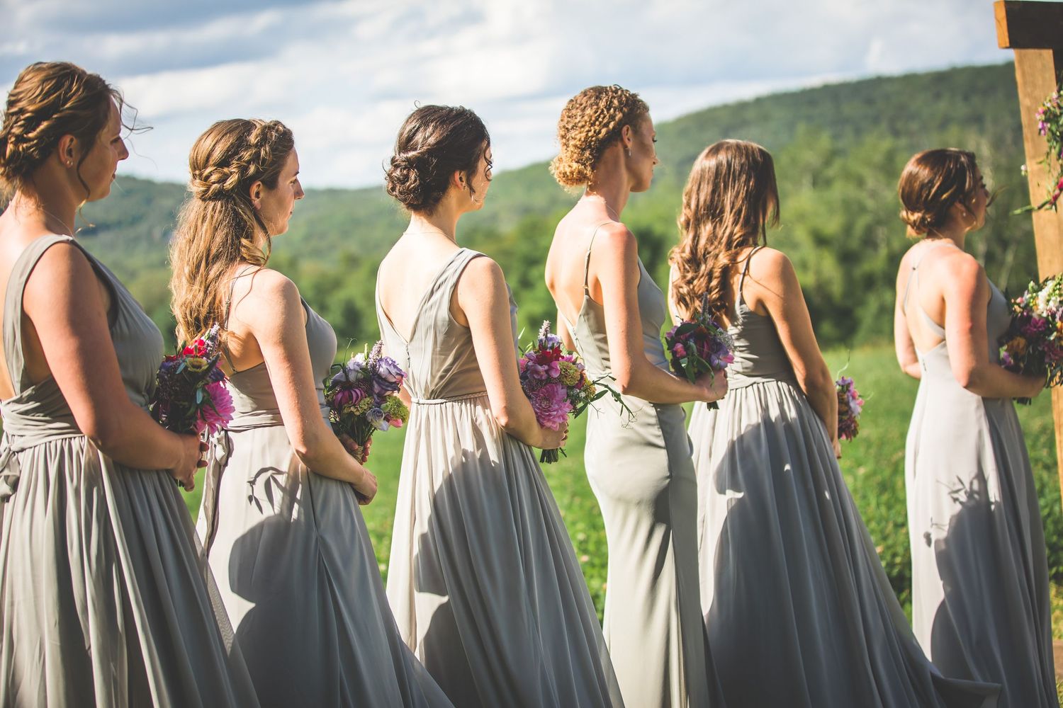 Row of bridesmaids in matching gray dresses standing on grassy hill during outdoor wedding.