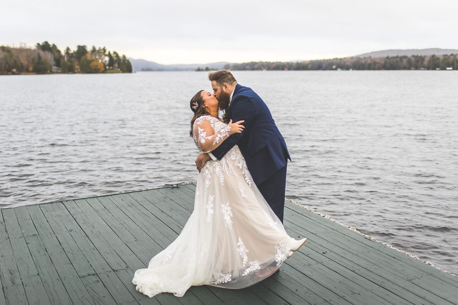 A romantic wedding moment captured on a lakeside dock with mountains in the background.