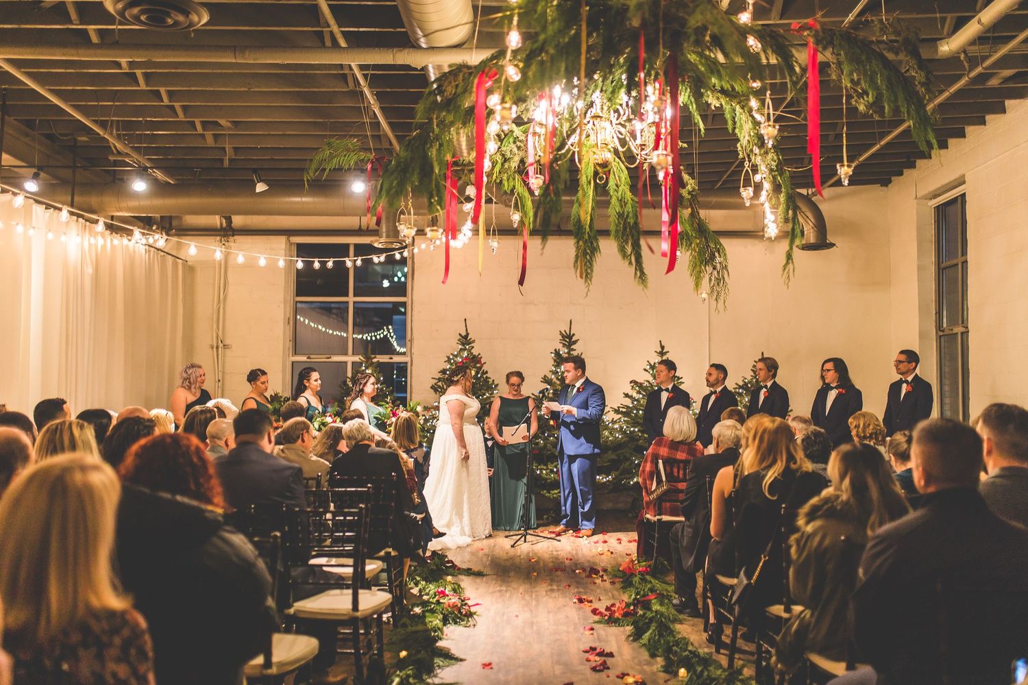 Wide view of wedding ceremony with guests seated in rows under decorative lighting and greenery.